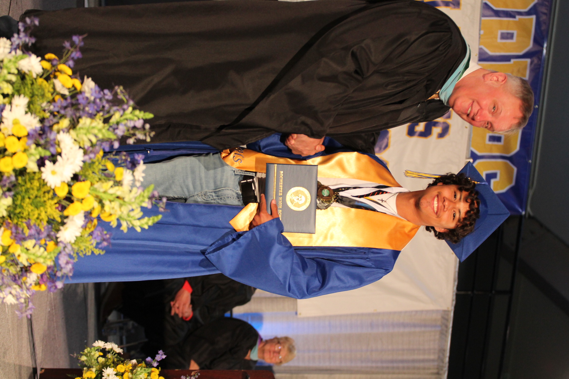 A male figure in a graduation gown and cap shakes hands with another individual. Both hold a certificate. Behind them, seated people watch the event.