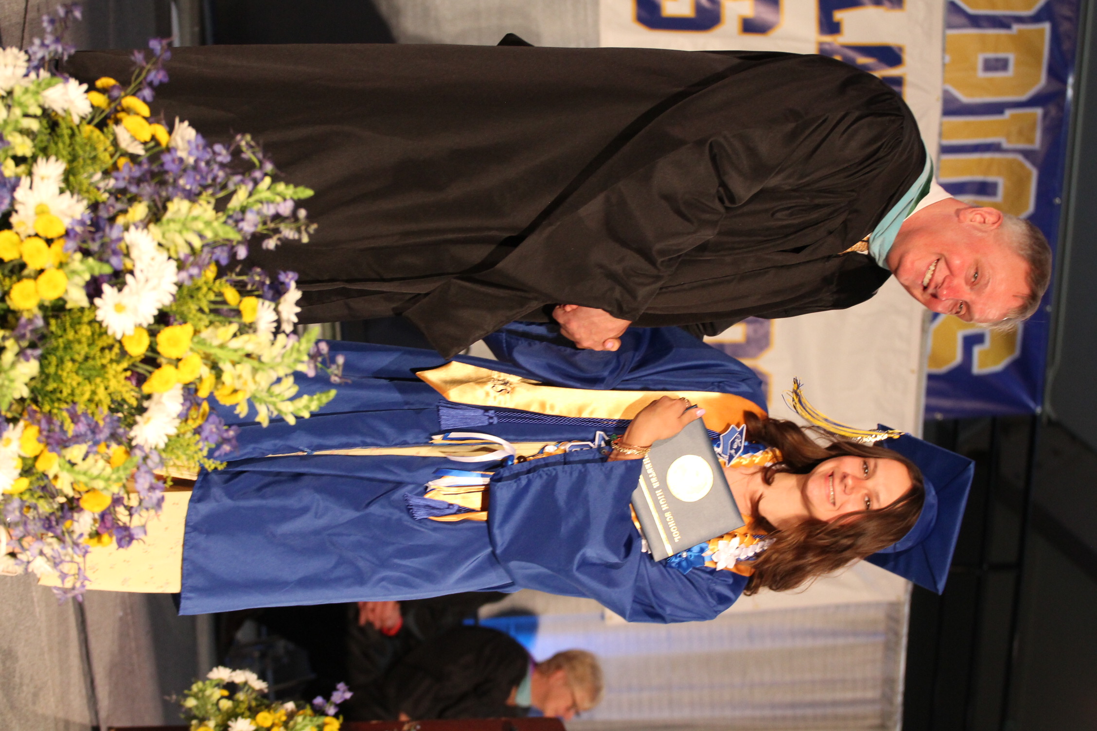 A male presenter in a black gown hands an award to a woman in a blue graduation gown on stage.