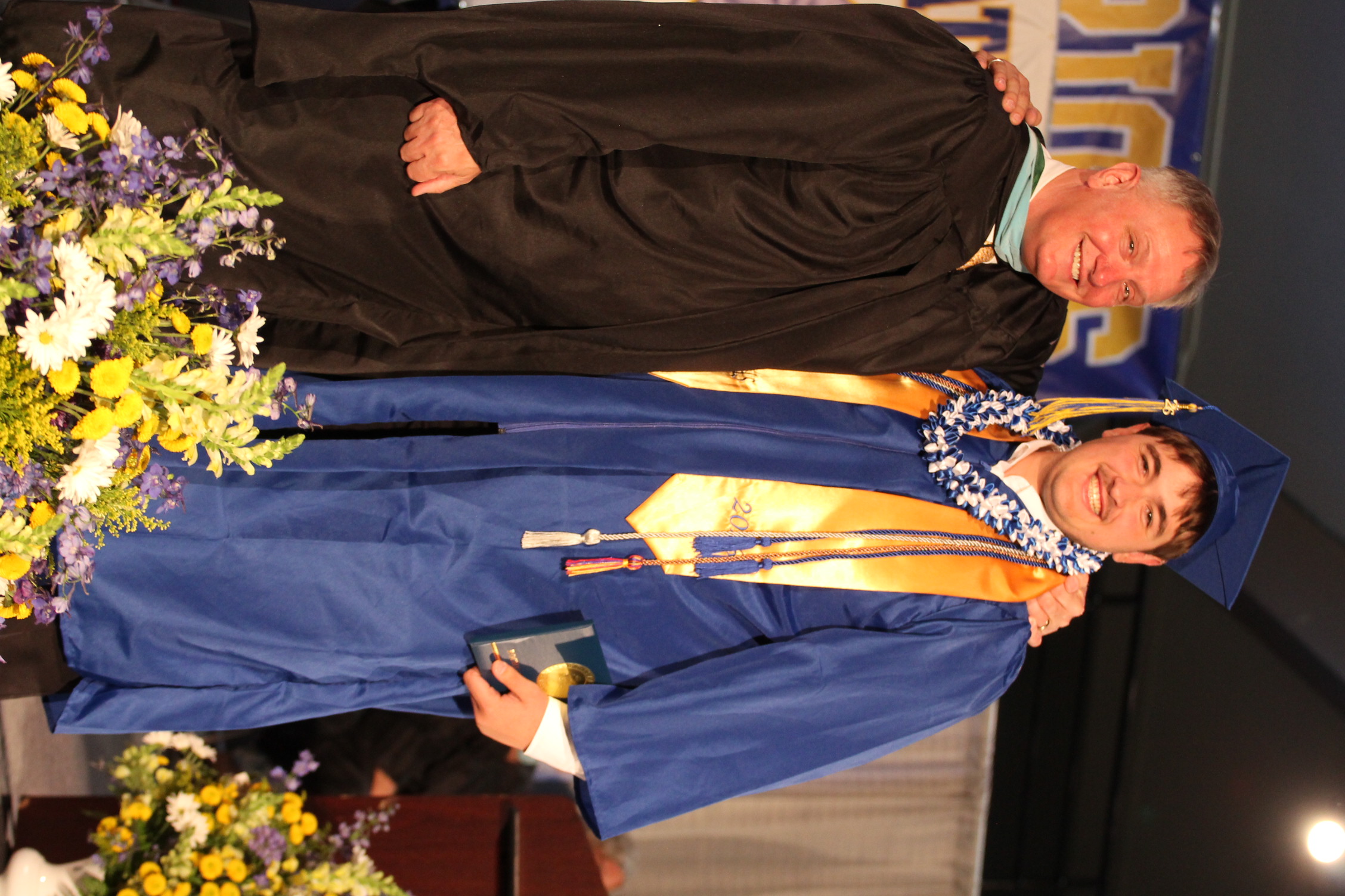 A man in a blue graduation gown with a gold stole stands next to a man in a black gown.