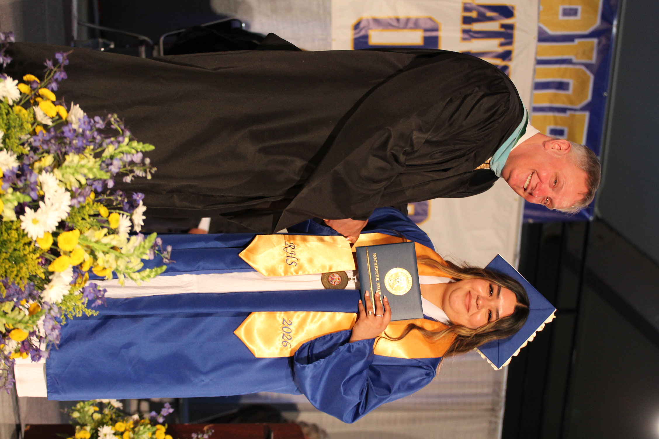 A graduate in a cap and gown holds a diploma, standing on stage with a smiling faculty member.