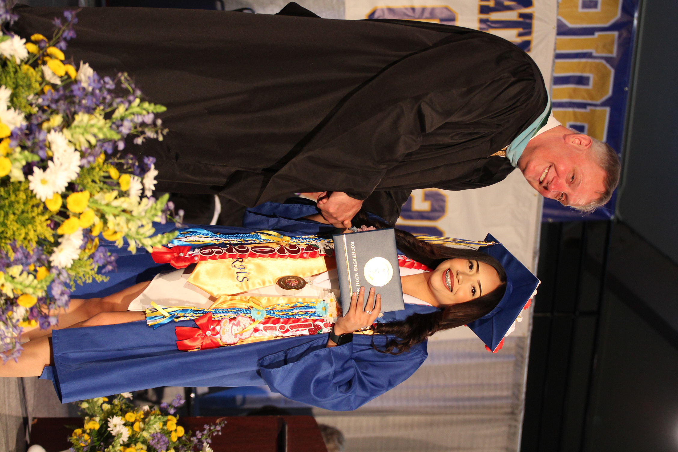 Man in black graduation gown giving diploma to a woman in blue gown with a cap.