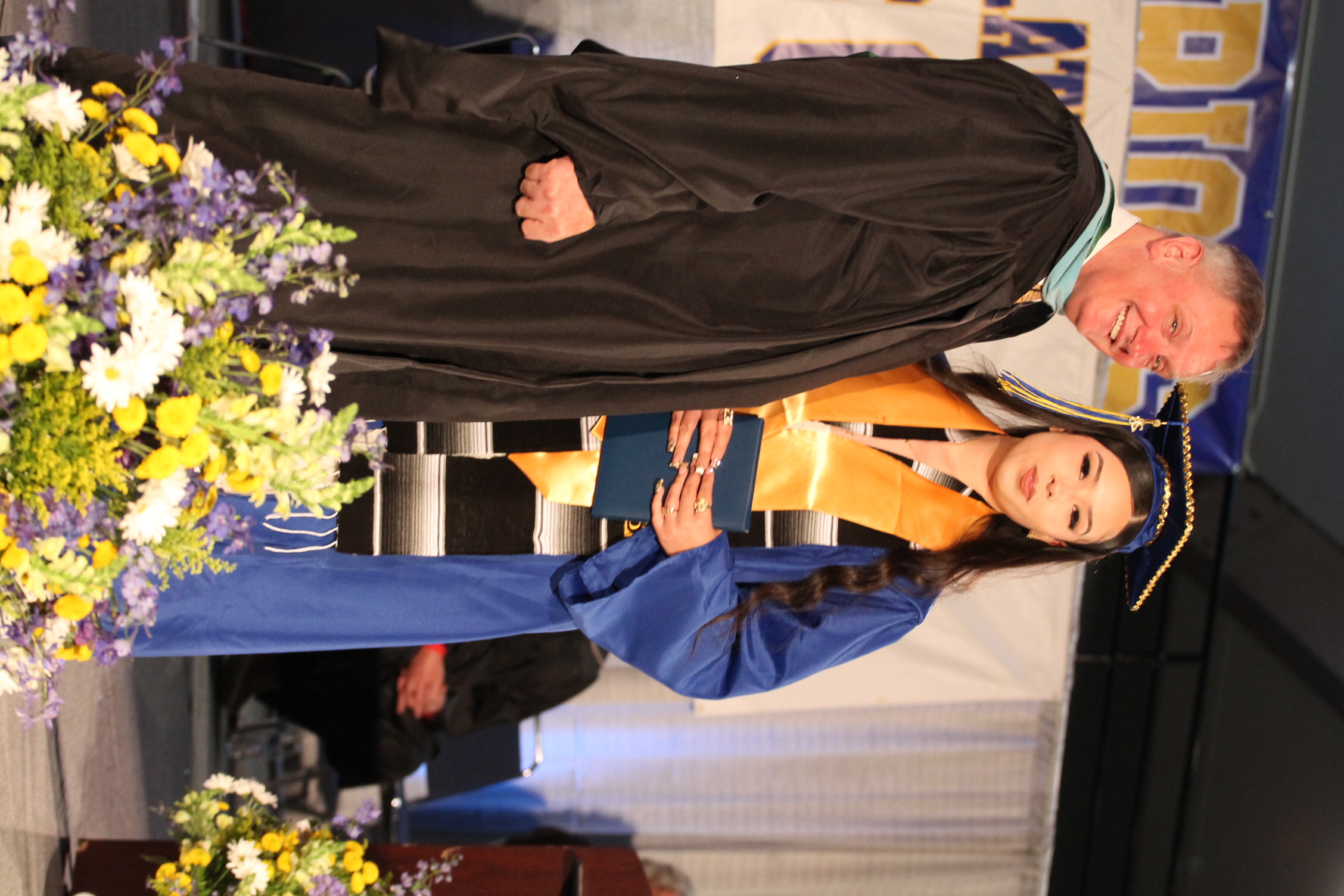 A man and woman on a stage, both dressed in academic gowns. The man is in black, the woman in blue.