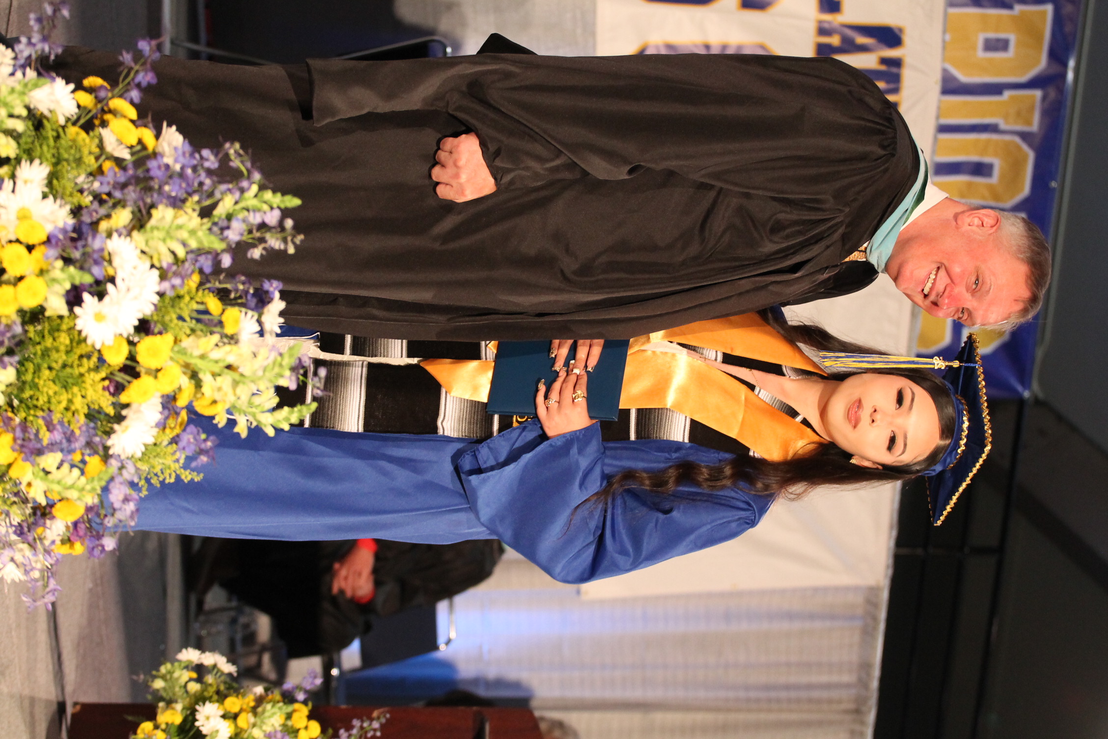 A man and woman in academic regalia stand on a stage. The woman holds a diploma. Floral arrangements are on the stage.
