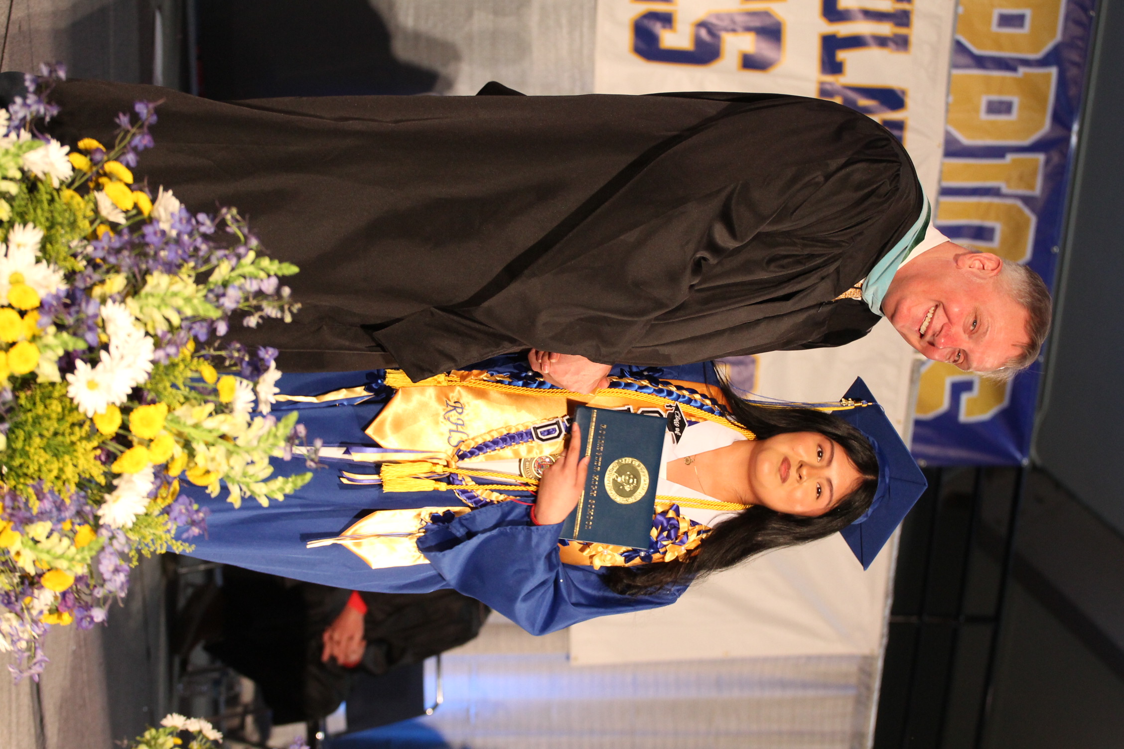 A man and woman stand on a stage. The man wears a black graduation gown, and the woman holds a certificate.