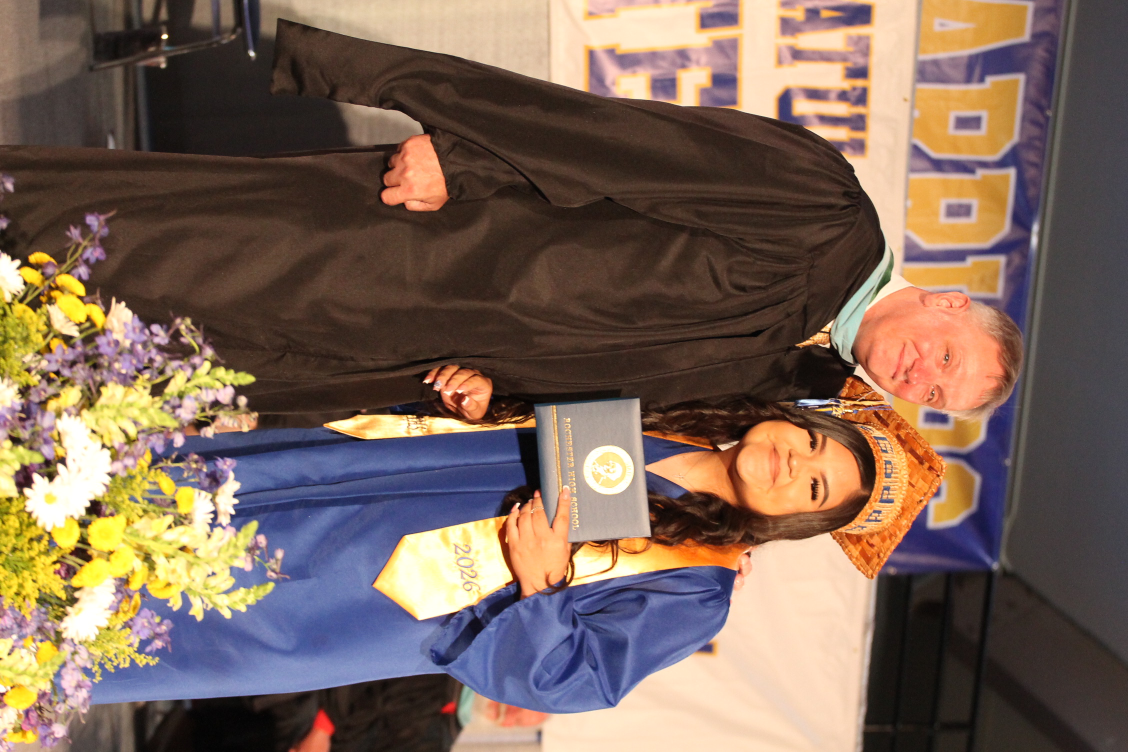 A man and woman stand on a stage at a graduation ceremony, with the woman holding an award.