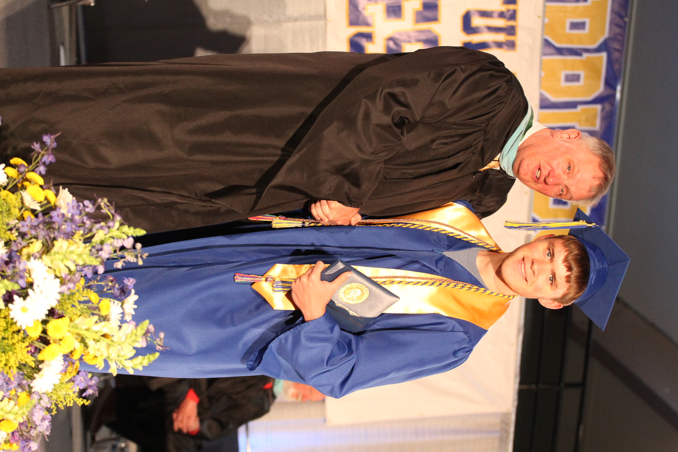 A man in a black gown stands beside a young man in a blue graduation gown. They both smile for the camera.