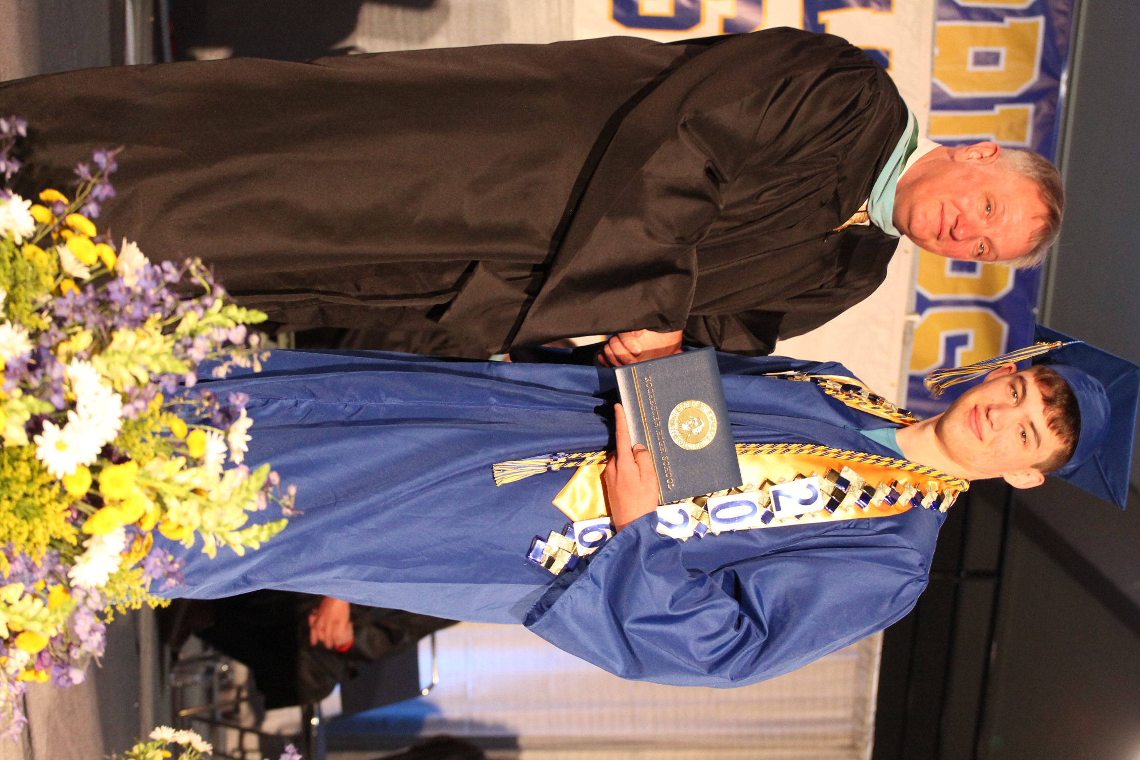 A man in a black gown hands a diploma to a young man in a blue gown. Both are standing on stage with flowers.