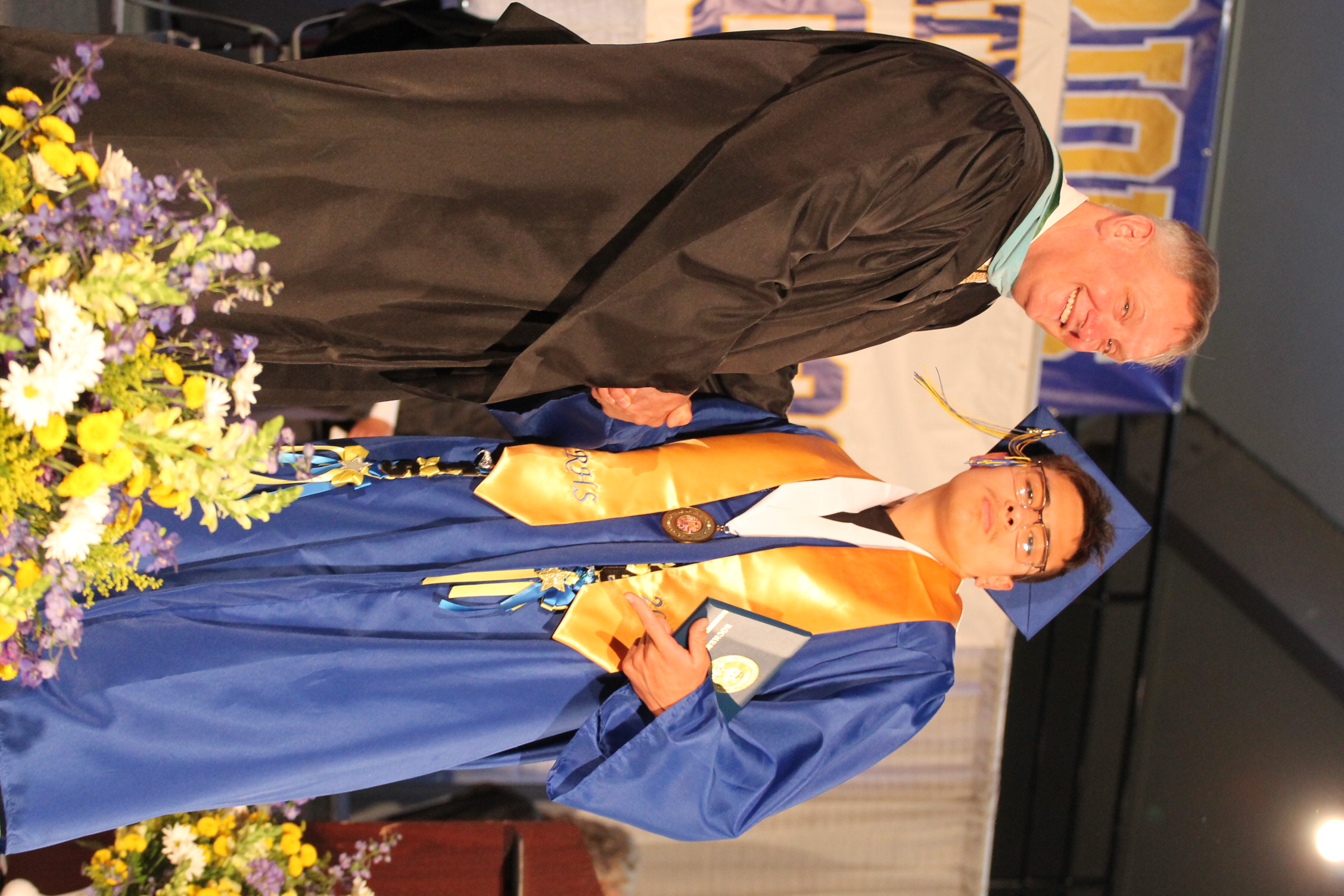 A graduate wearing a blue gown and glasses stands next to a man in a black gown at a ceremony.