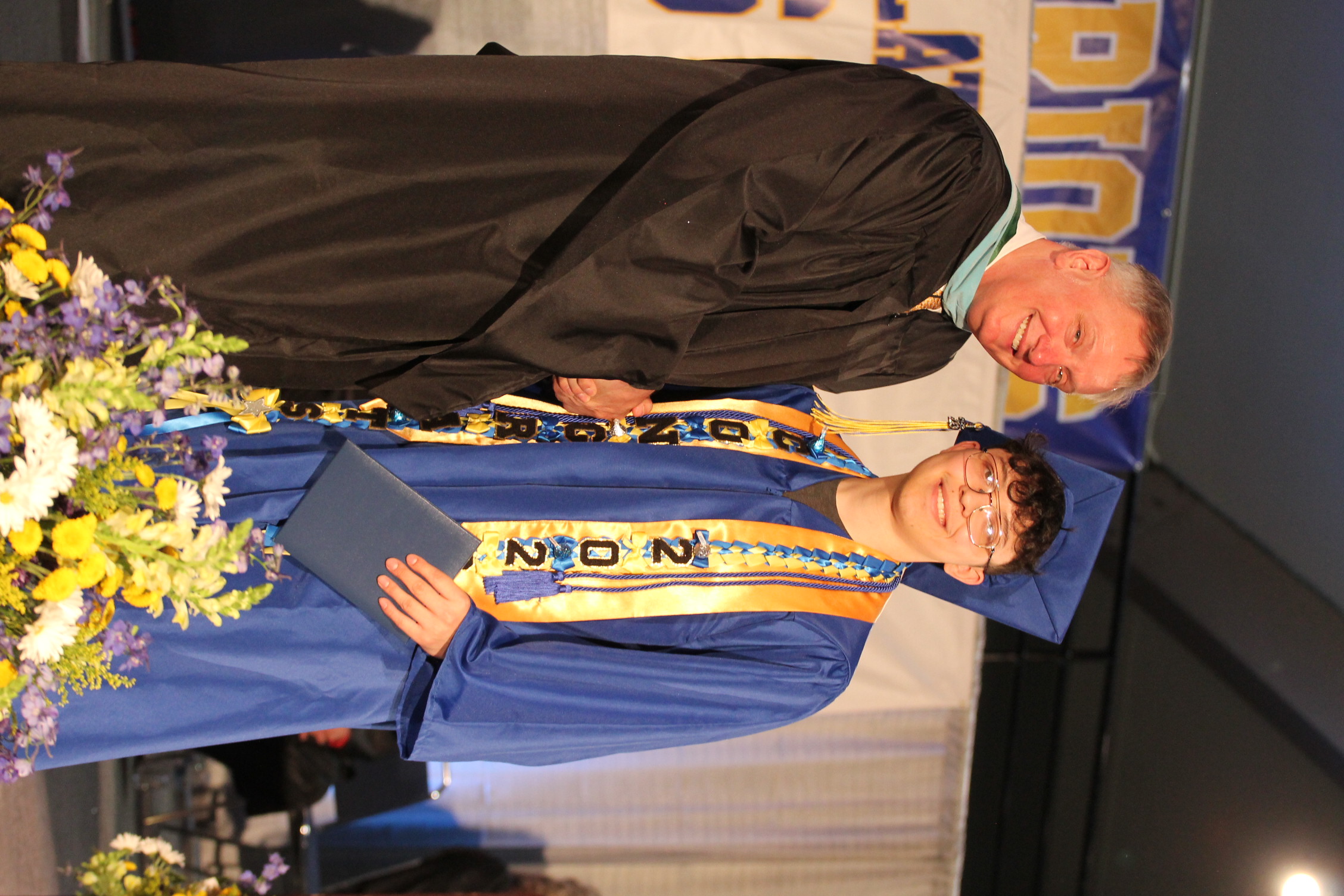 A man in a black robe and a youth in a blue gown stand next to each other, smiling. A banner behind them says "DINING."