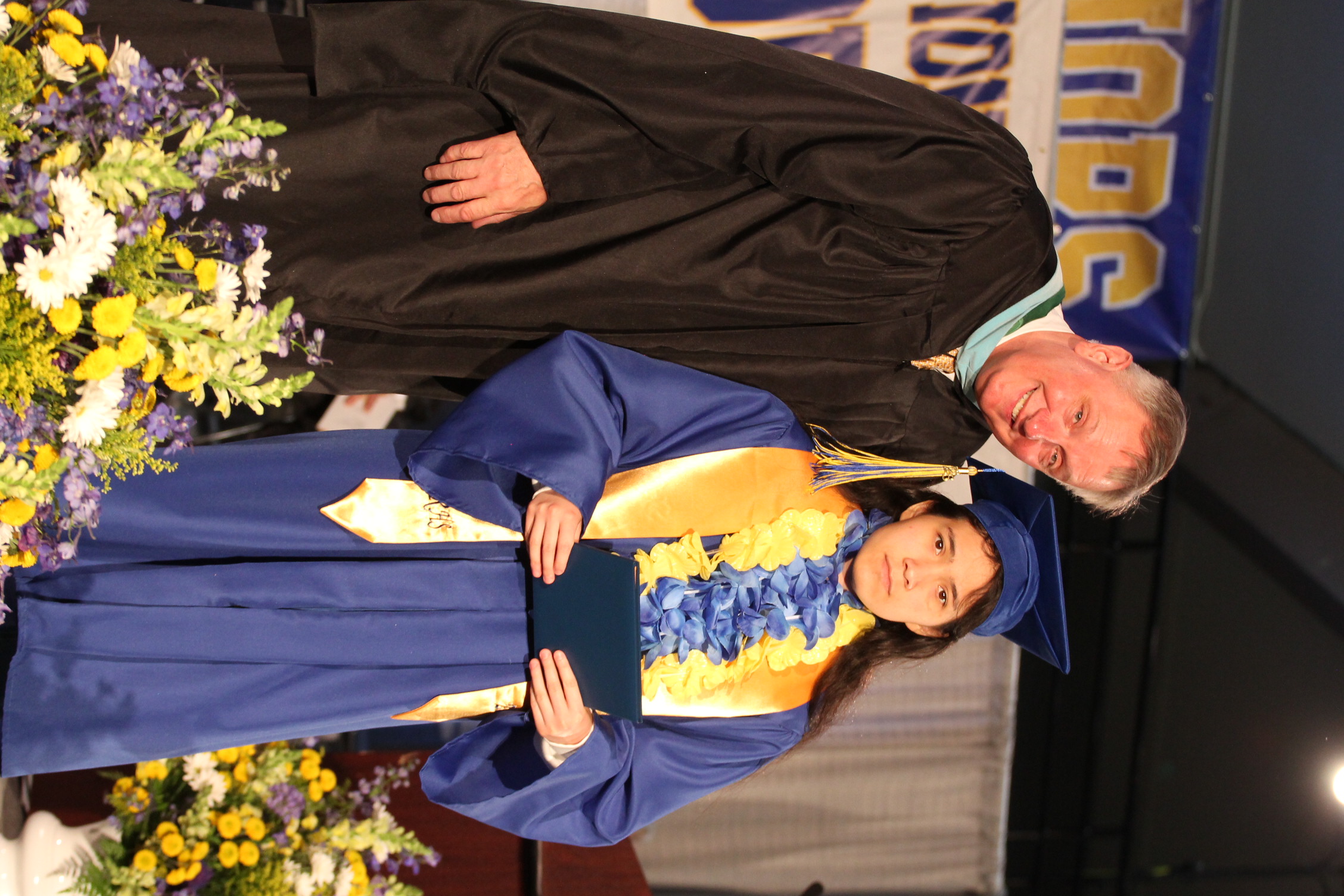 A man in a black robe and a young girl in a blue gown stand at a graduation ceremony.