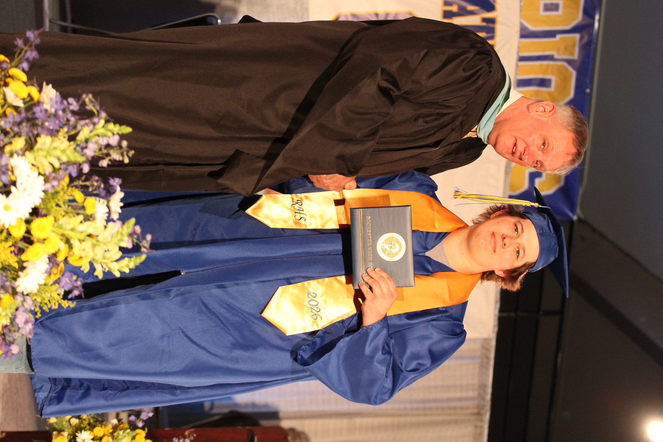 A male adult in a black graduation gown hands a certificate to a woman in a blue gown.