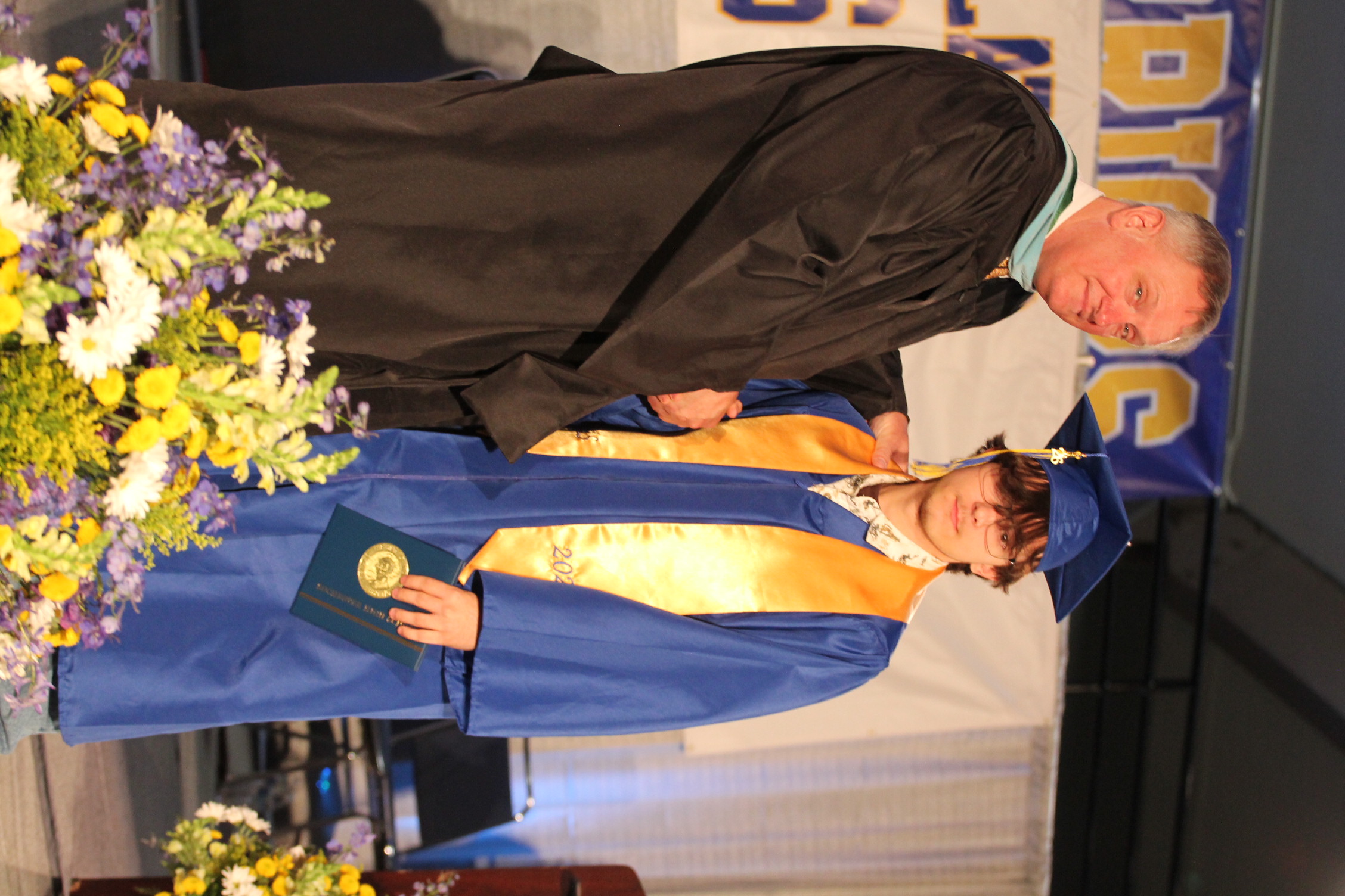 A man in a black robe and a young person in a blue gown and cap shake hands.