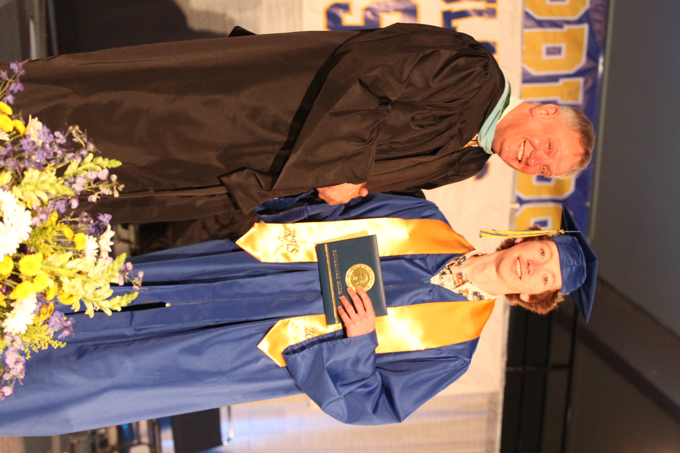 A man in a black robe and a woman in a blue gown with a gold stole shake hands on stage. A banner and flowers are in the background.
