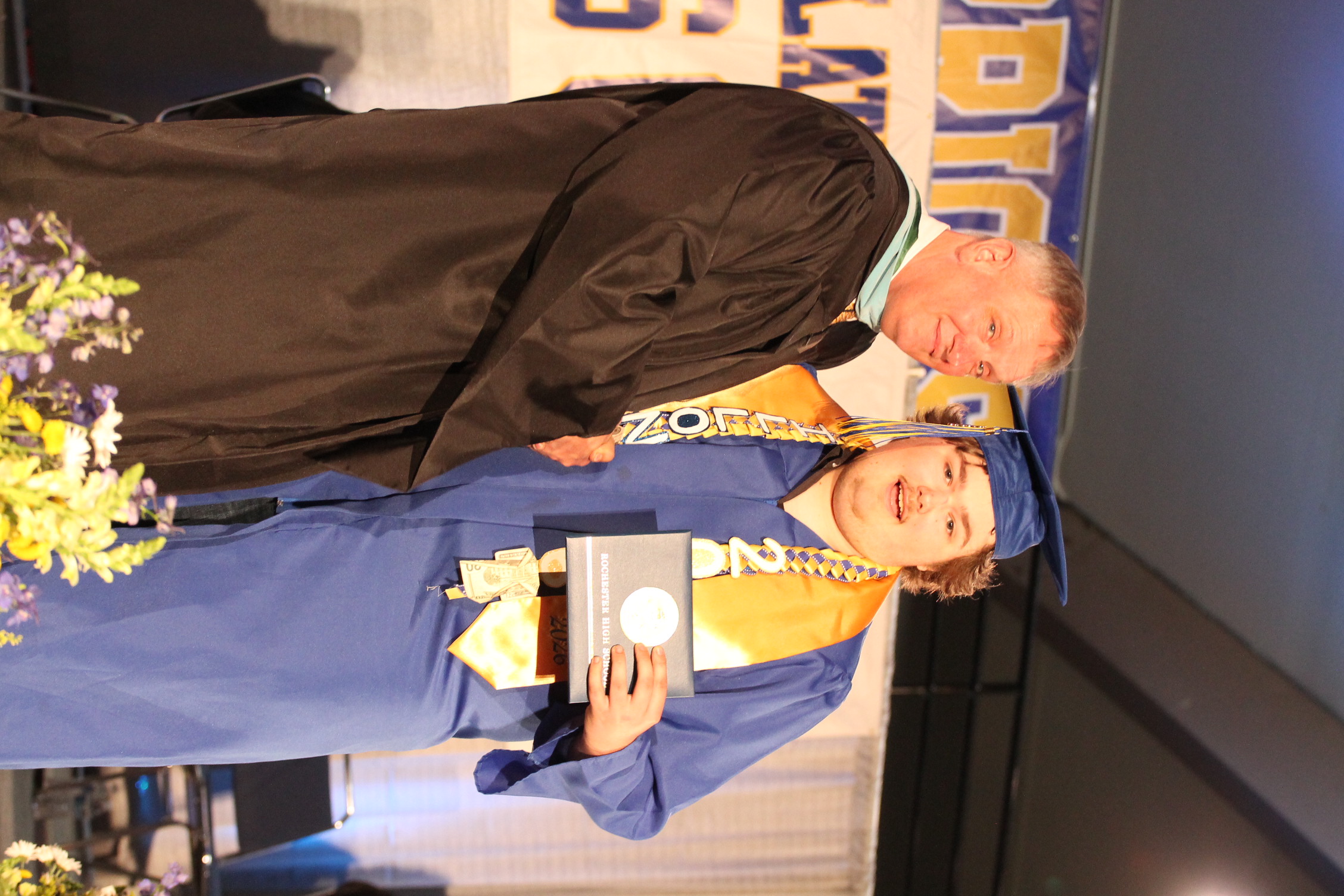 A man in a brown gown hands a diploma to a graduate wearing a blue gown and cap.