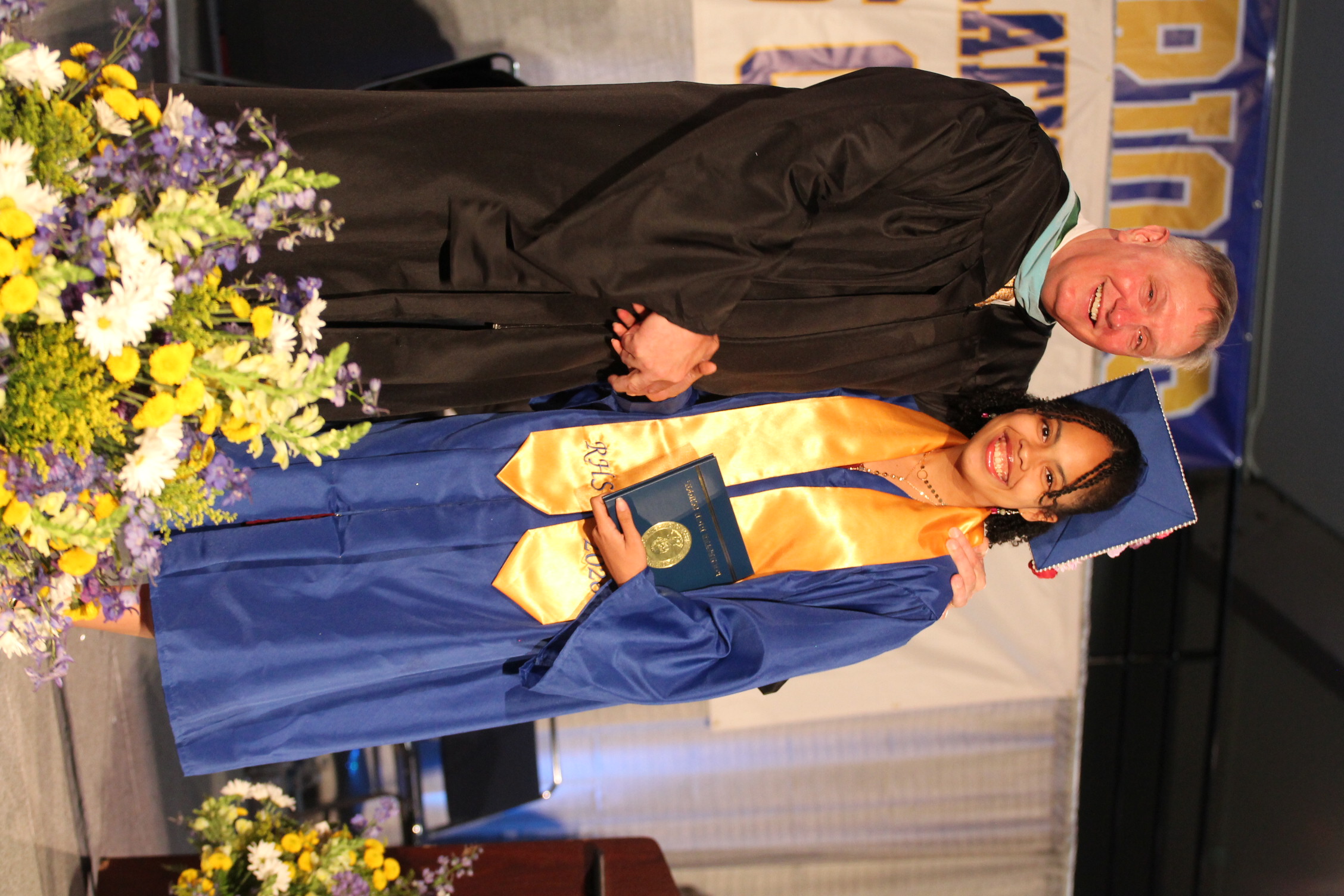 A male faculty member and a female graduate, both wearing gowns, stand together on stage, holding hands.
