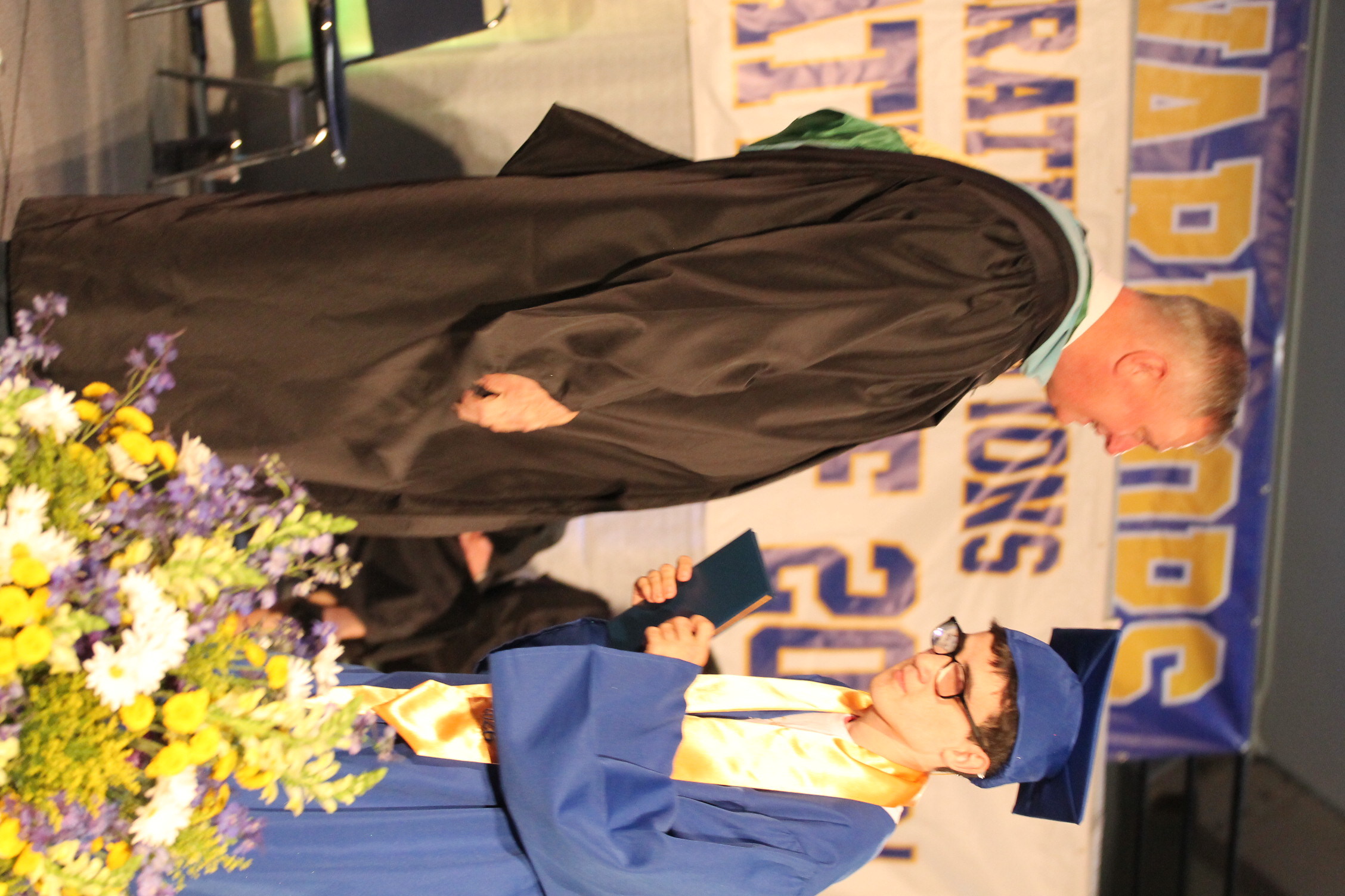 Two men in academic regalia on stage. One holds a diploma and the other wears a graduation cap.