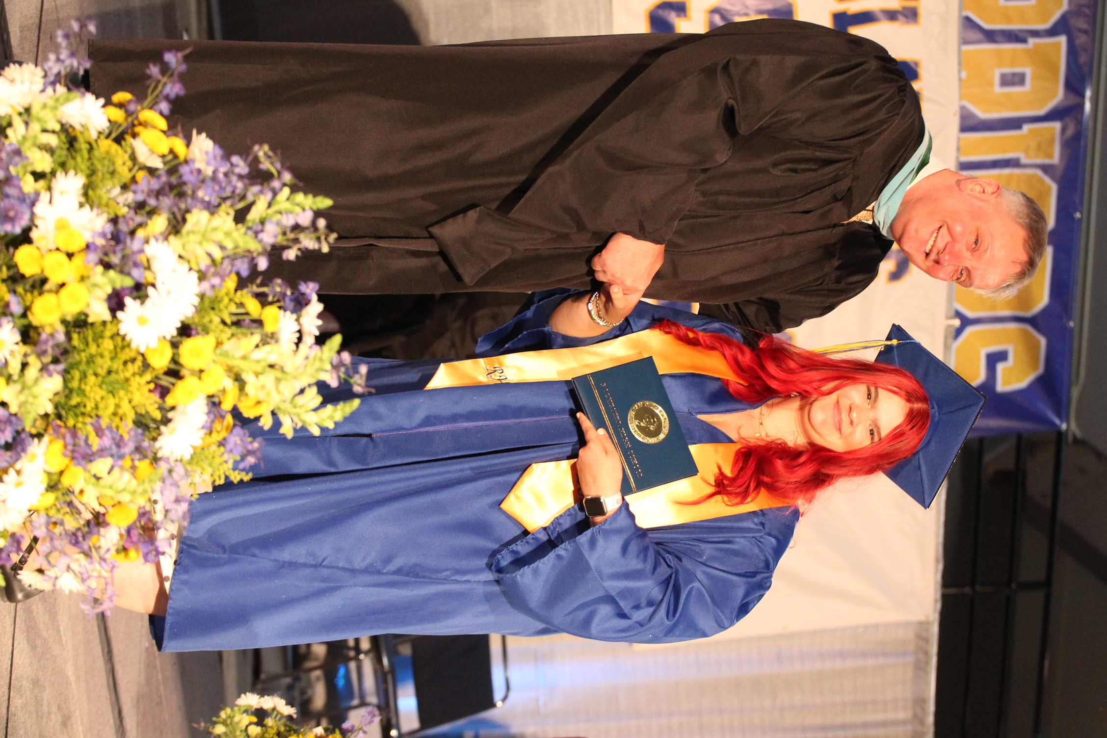 A graduate, wearing a blue gown and red hair, accepts a diploma from a faculty member. They stand on stage with a backdrop and floral arrangements.