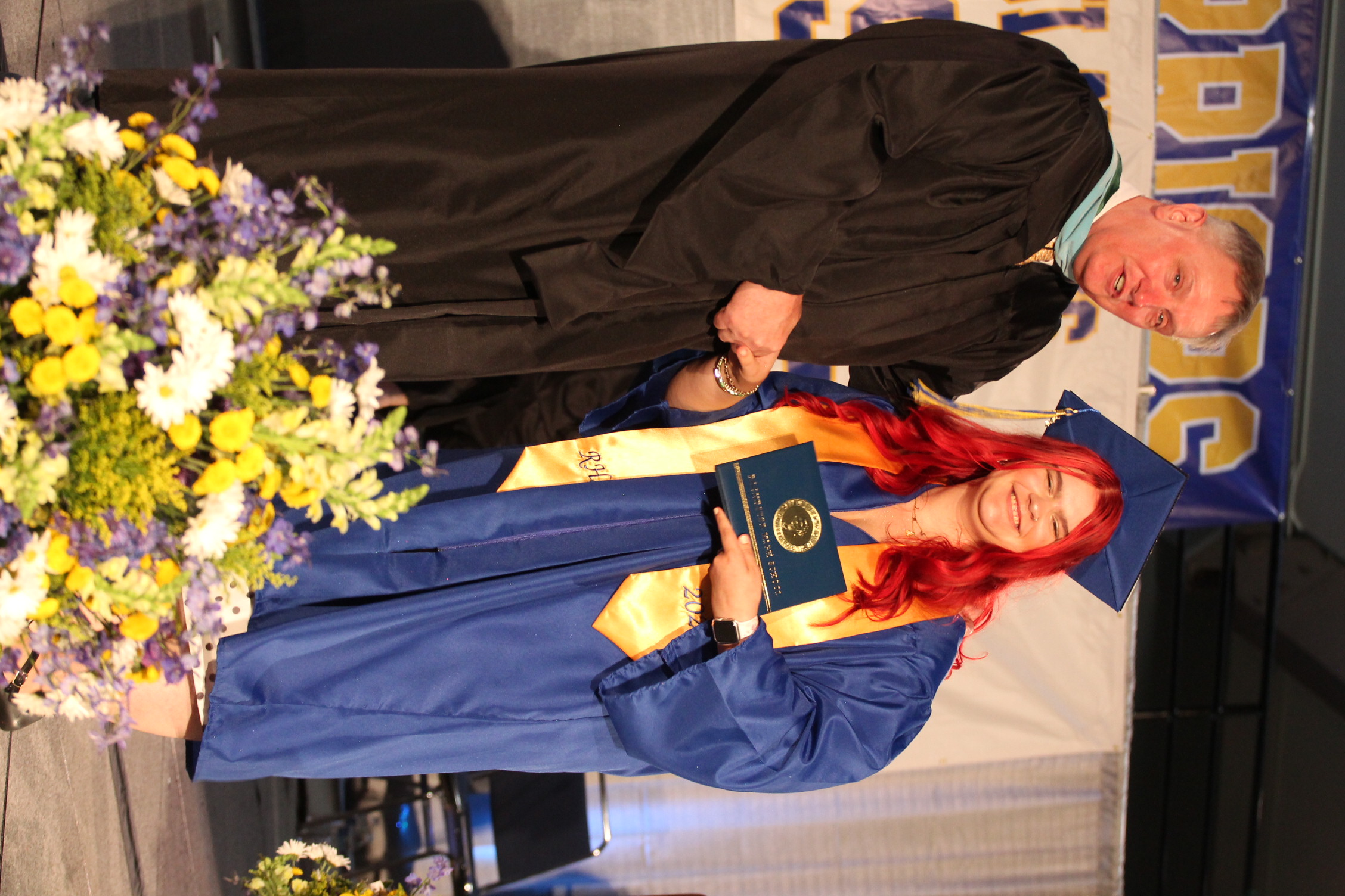 A graduate in blue gown holds a diploma as a presenter in black gown hands it to them on stage.