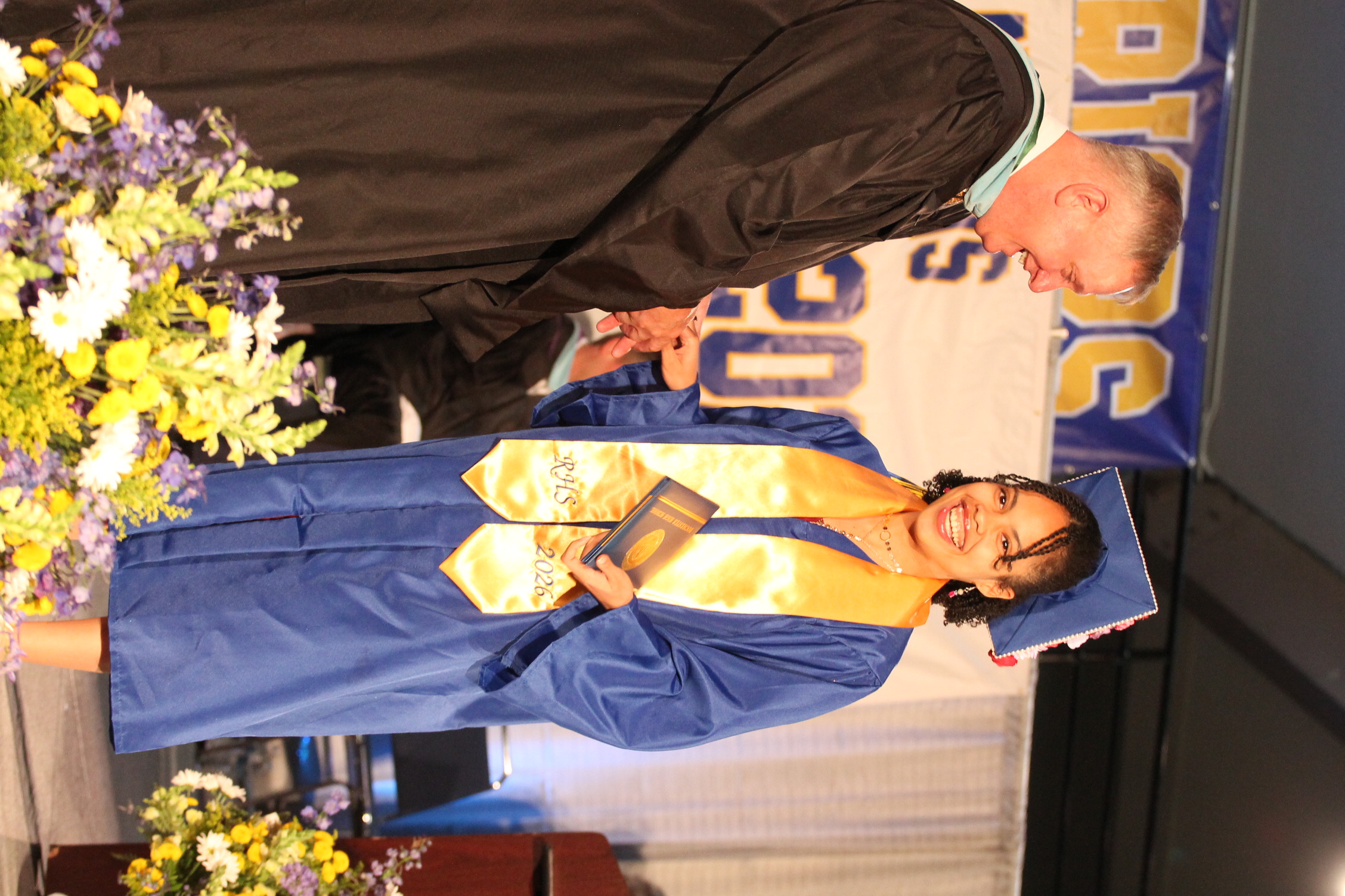 A male in a black graduation robe hands a diploma to a female in a blue gown.