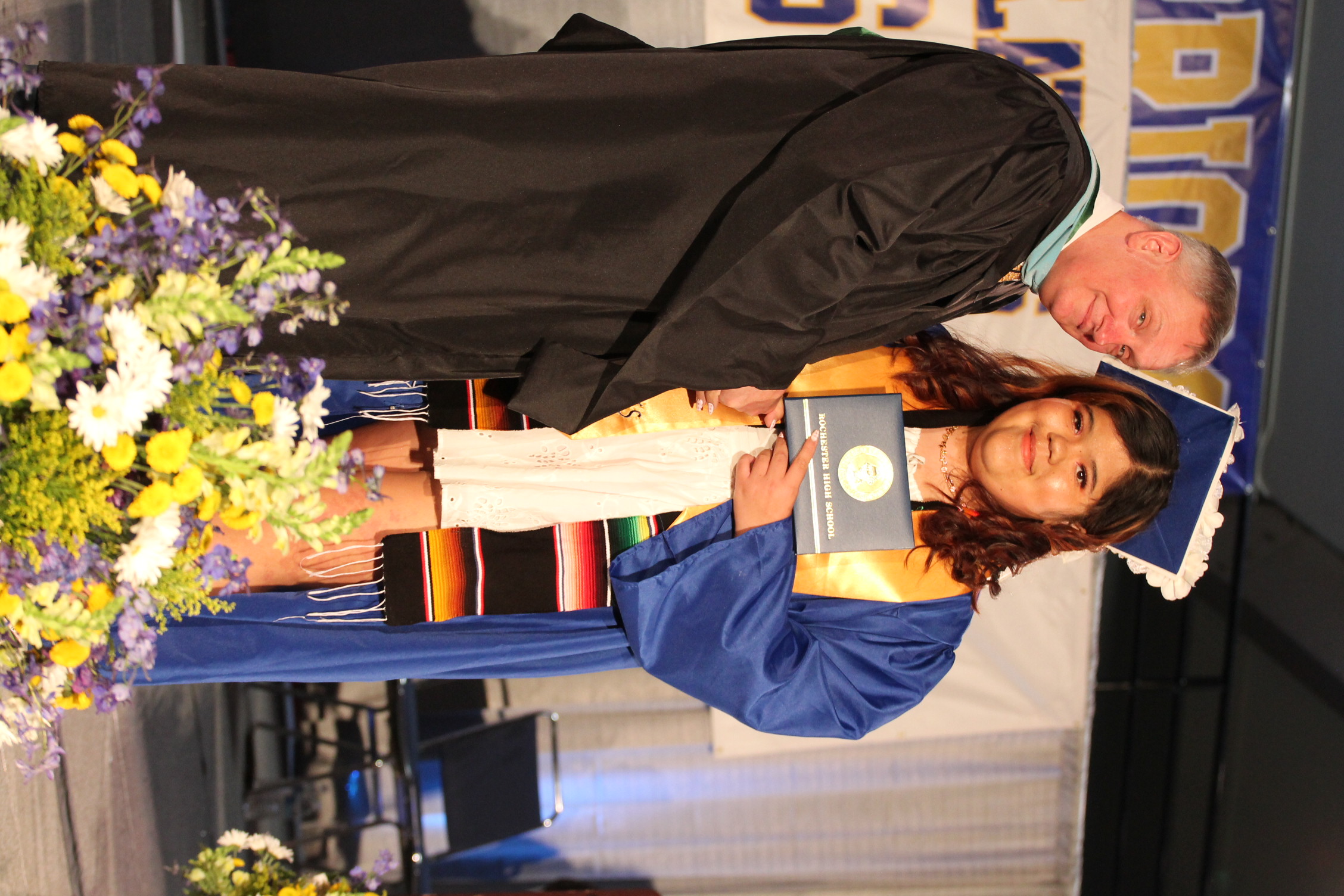 A graduate in a blue gown and a male faculty member in a black gown hold an award.
