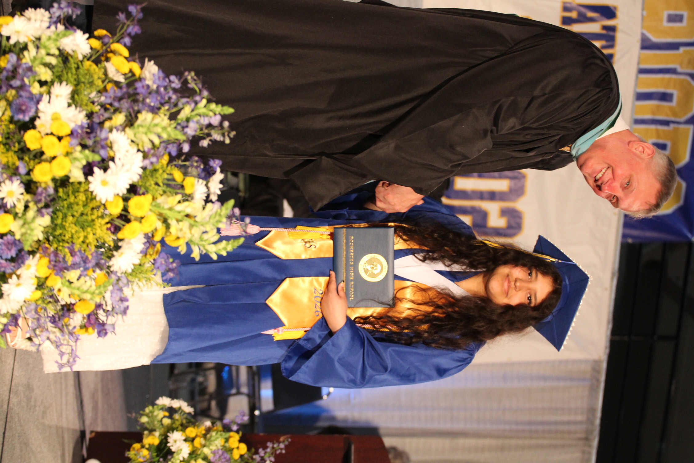 A man in a black gown shakes hands with a woman in a blue gown holding a framed award.