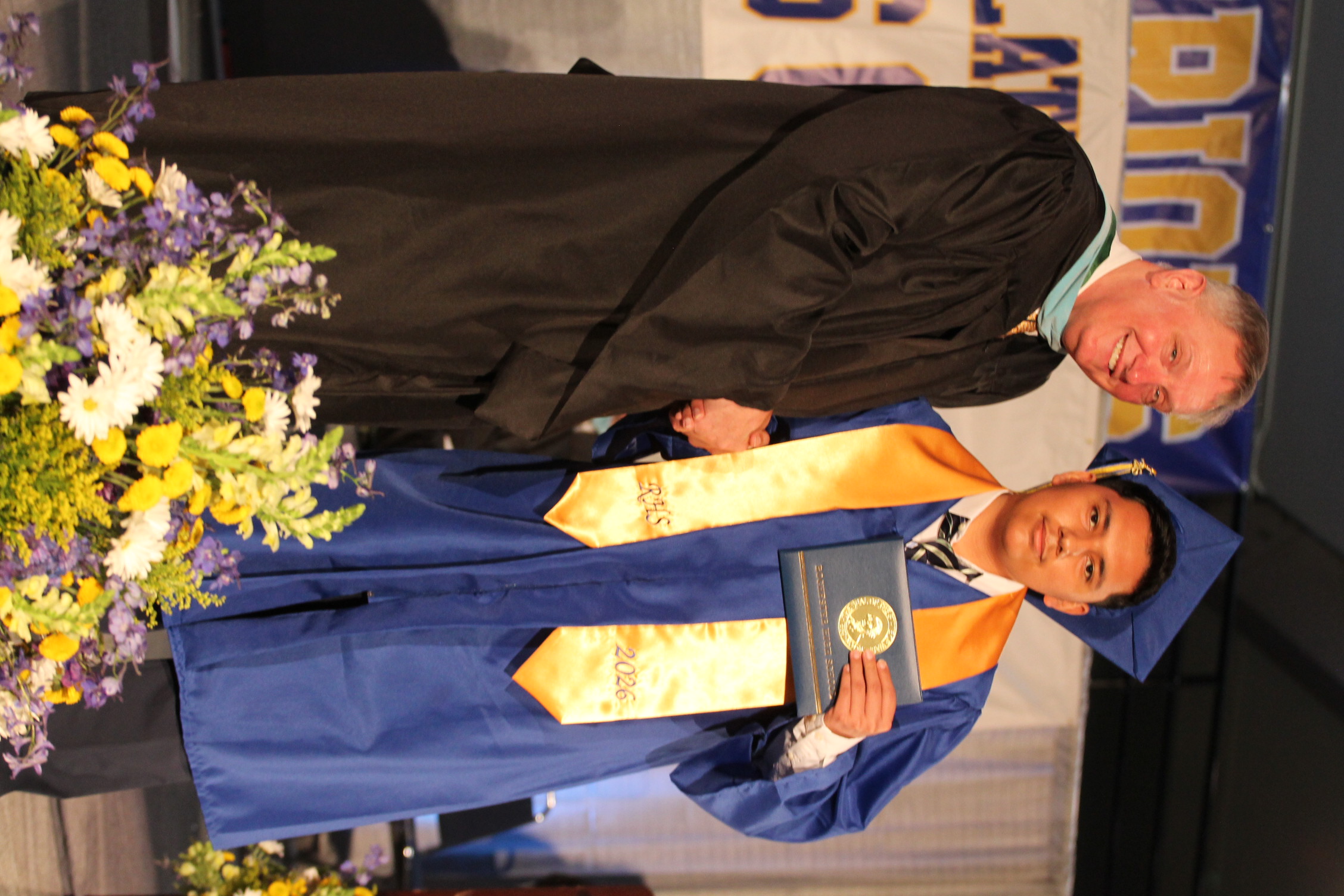 A man in a black graduation gown shakes hands with a graduate wearing a blue gown. A large flower arrangement is in the foreground.