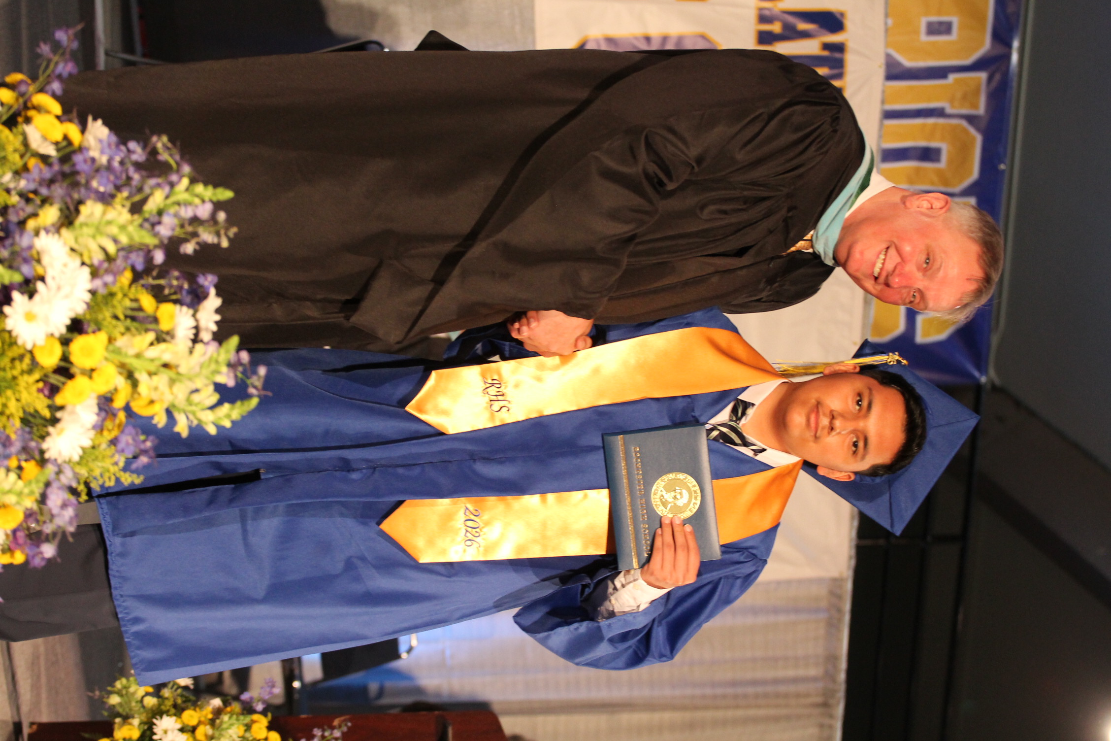 A man in a black robe and another in a blue robe with a graduation cap shake hands. One holds a diploma. Behind them, a banner reads "DISTRICT".