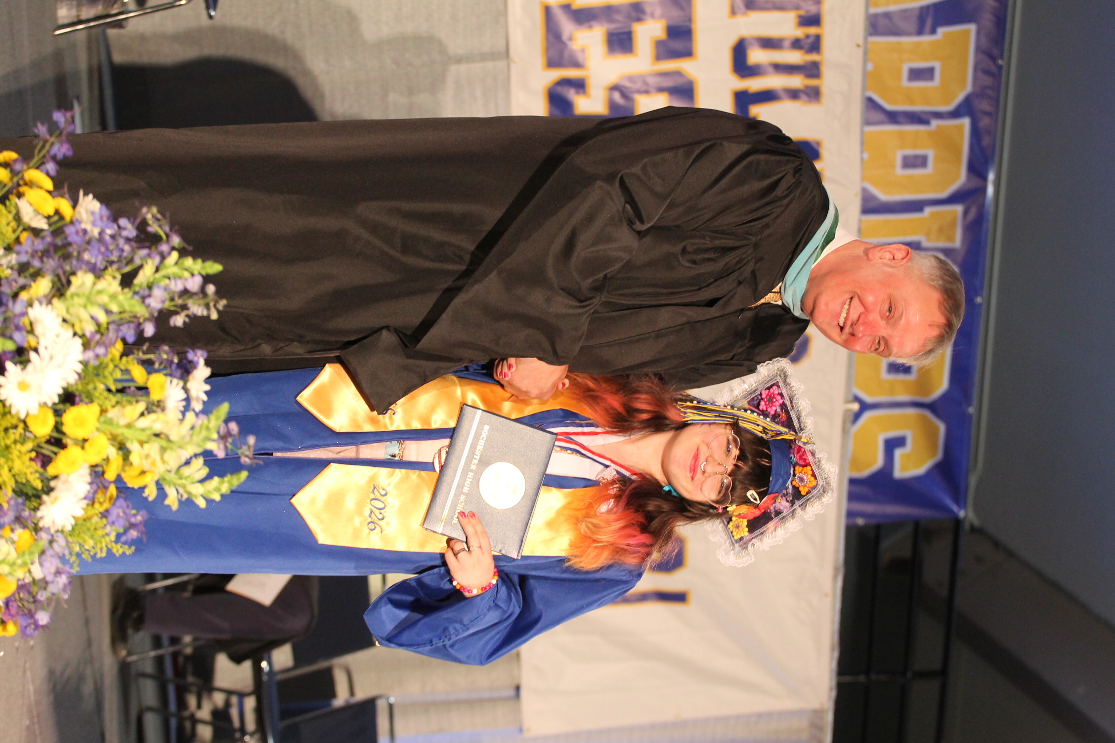 A man and a woman in graduation gowns on stage. The woman holds a diploma. A banner and flowers are in the background.
