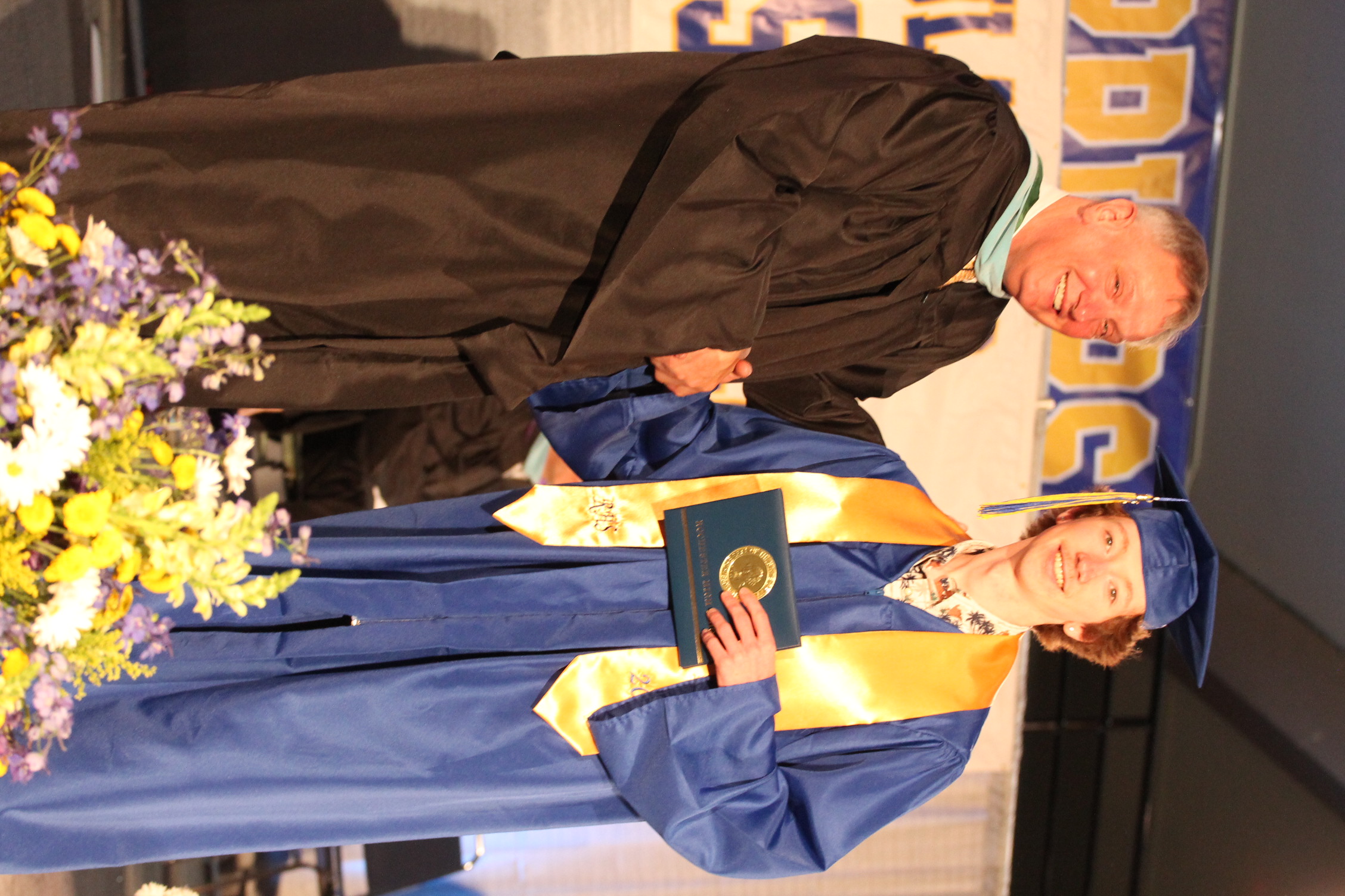 A person in a brown graduation gown shakes hands with a person in a blue gown, both smiling. The blue gown holder holds a certificate.
