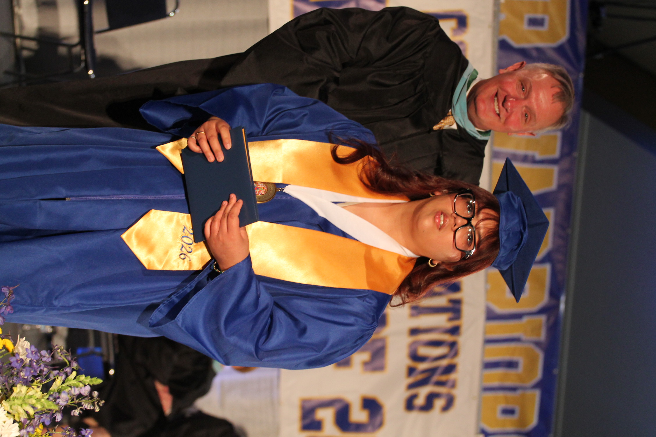 A person in a blue gown holds a diploma while standing on stage in front of a banner.