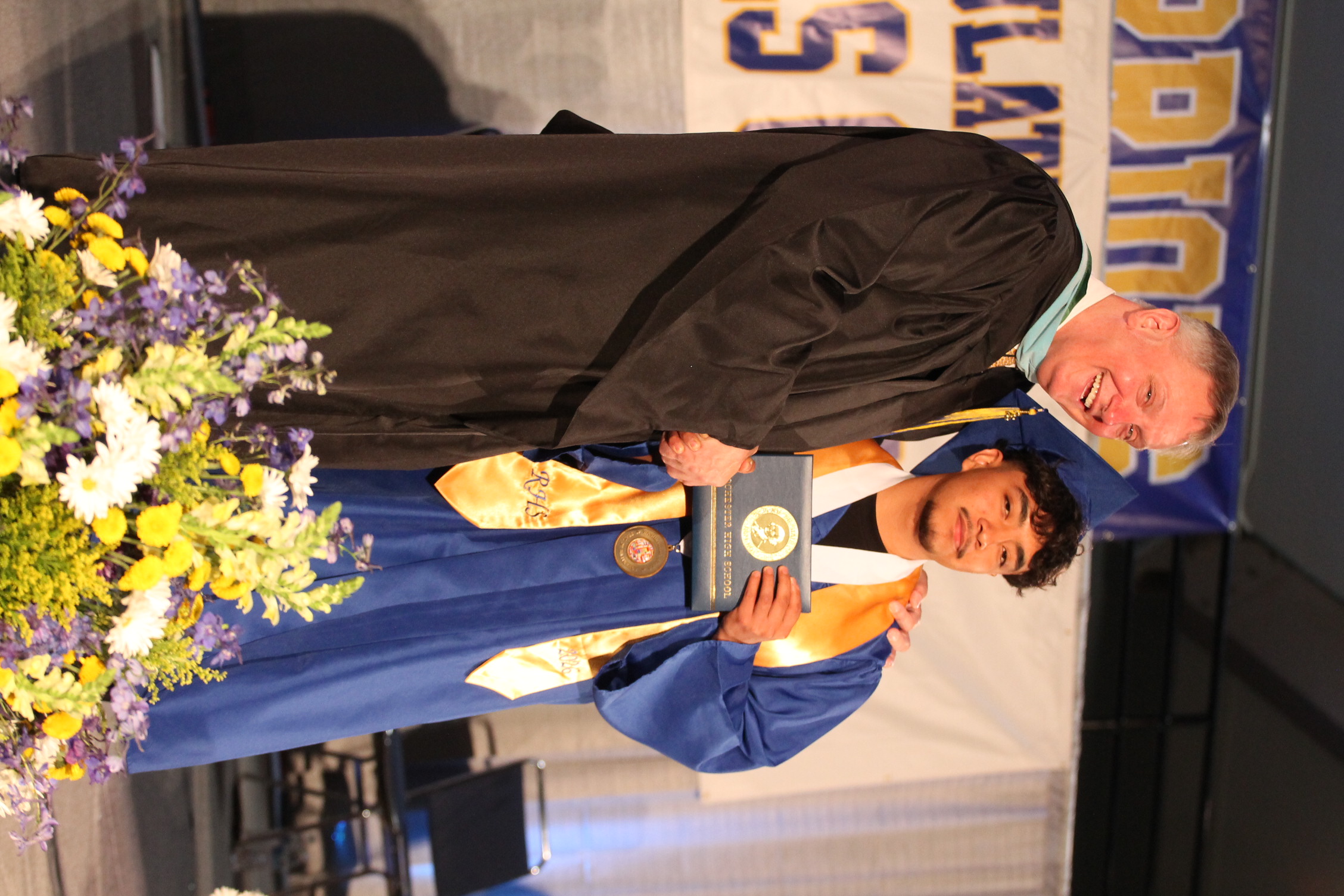 A man in a black gown, likely a teacher, presents an award to a graduate in a blue gown.