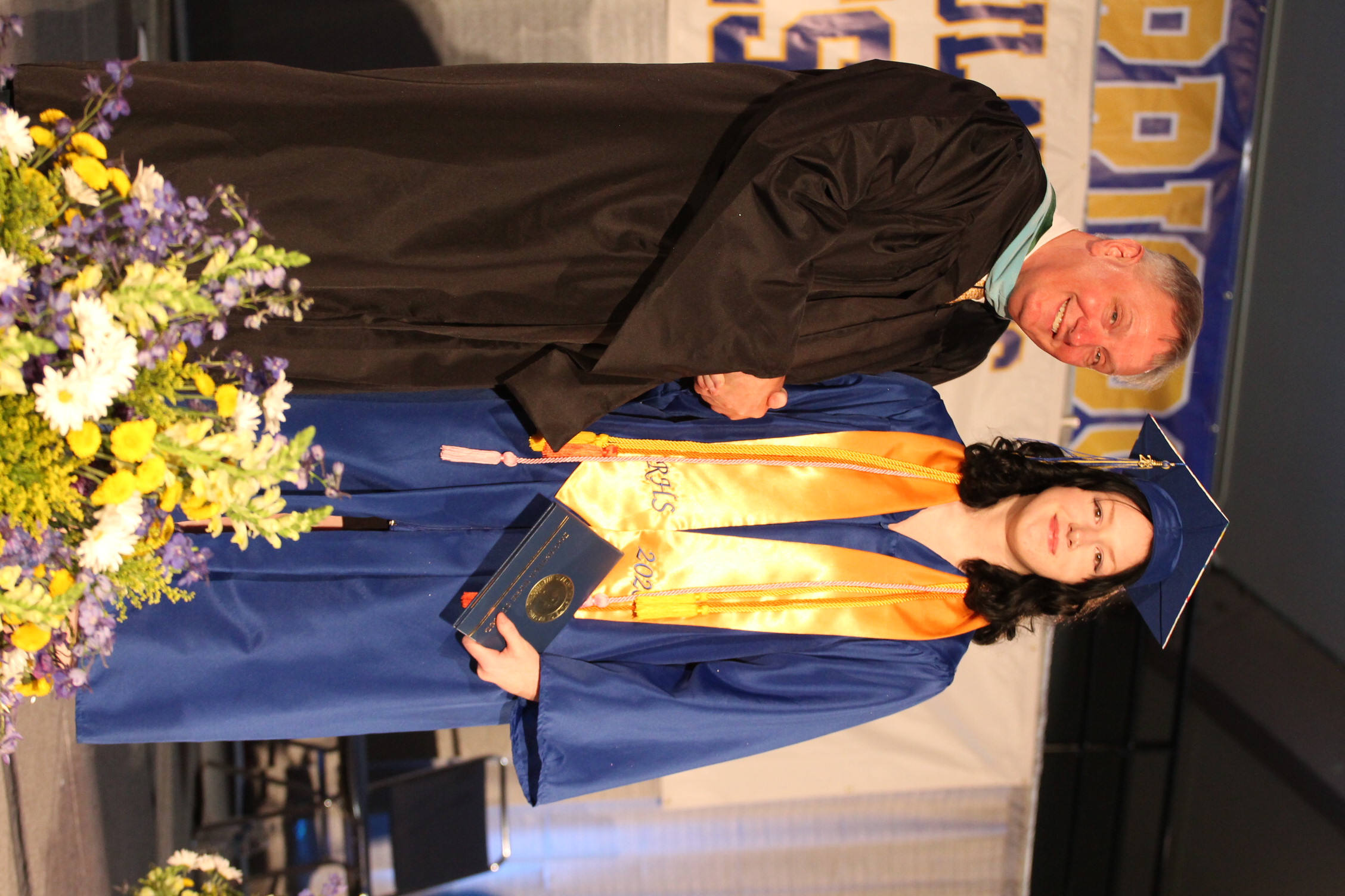 A man and a woman in academic regalia stand together. The man shakes the woman's hand. A bouquet of flowers is in the foreground.