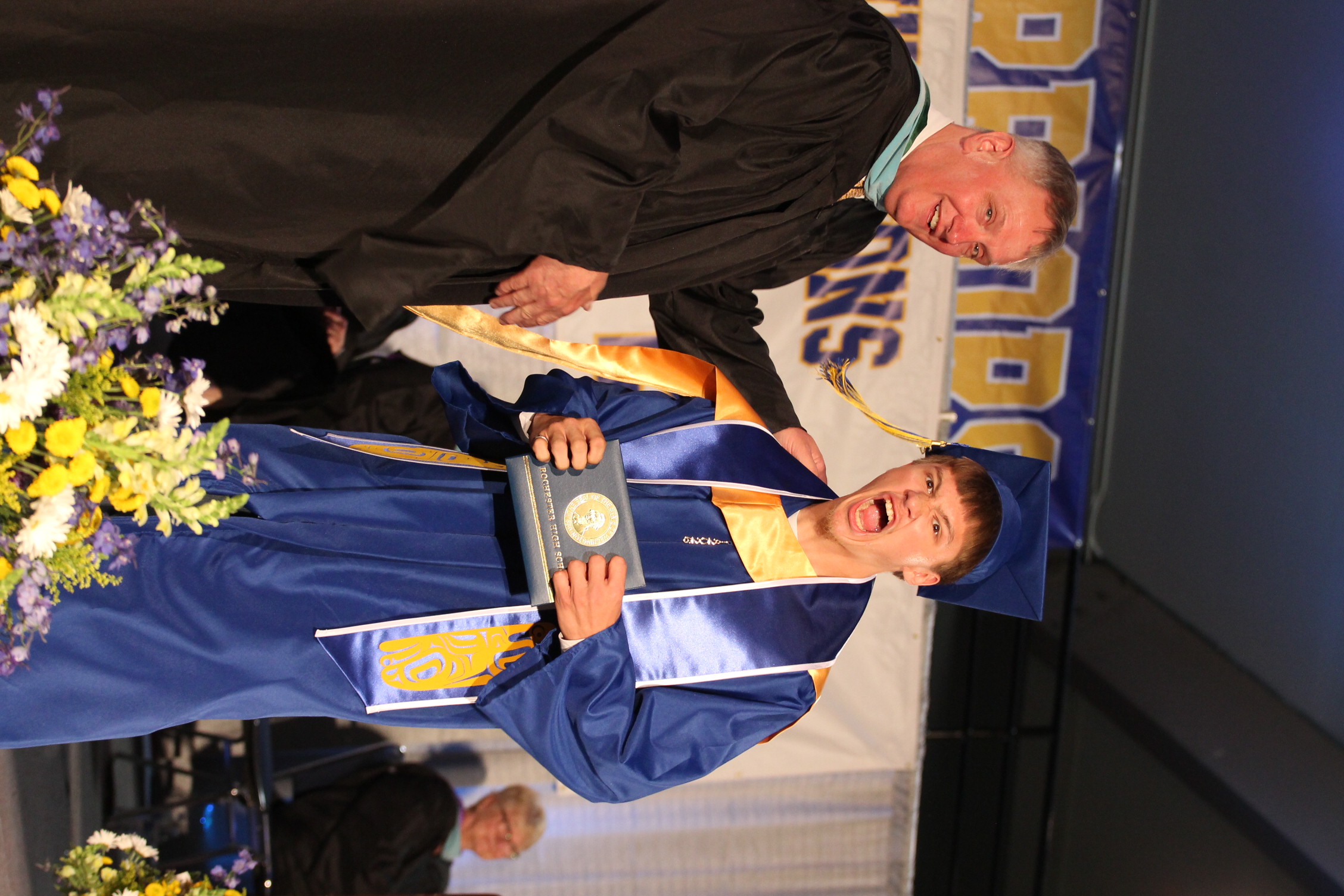 A man in a black robe hands a plaque to a young man in a blue cap and gown.