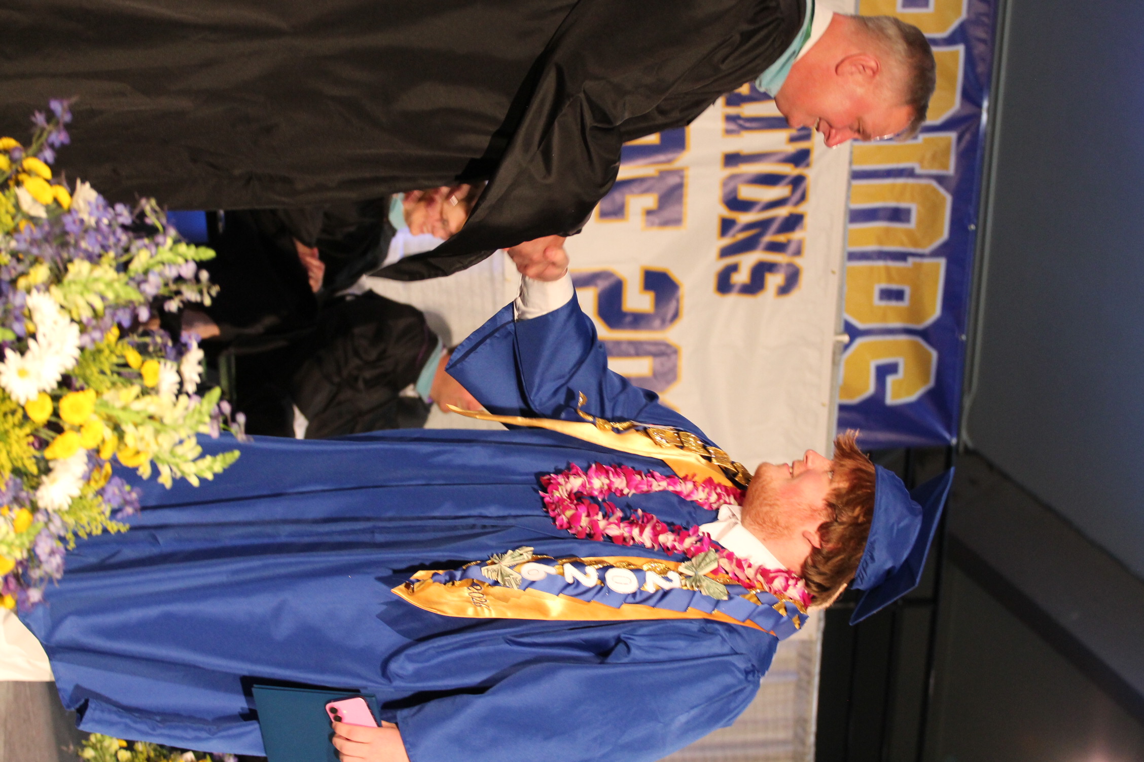 Man in black gown shakes hands with a man in blue gown and lei in front of audience.