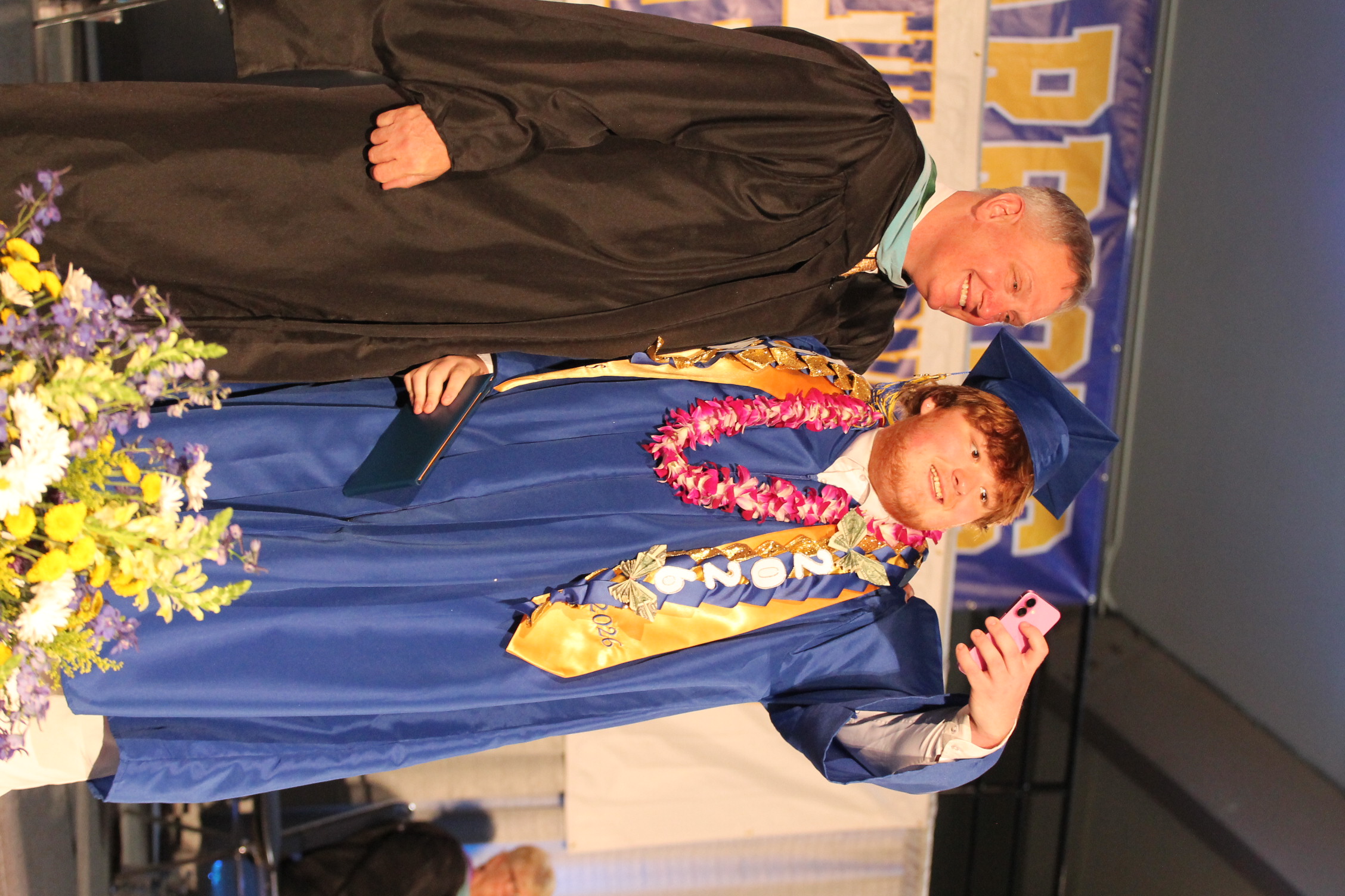 A man in a black gown and a woman in a blue gown stand next to each other with flowers on stage.