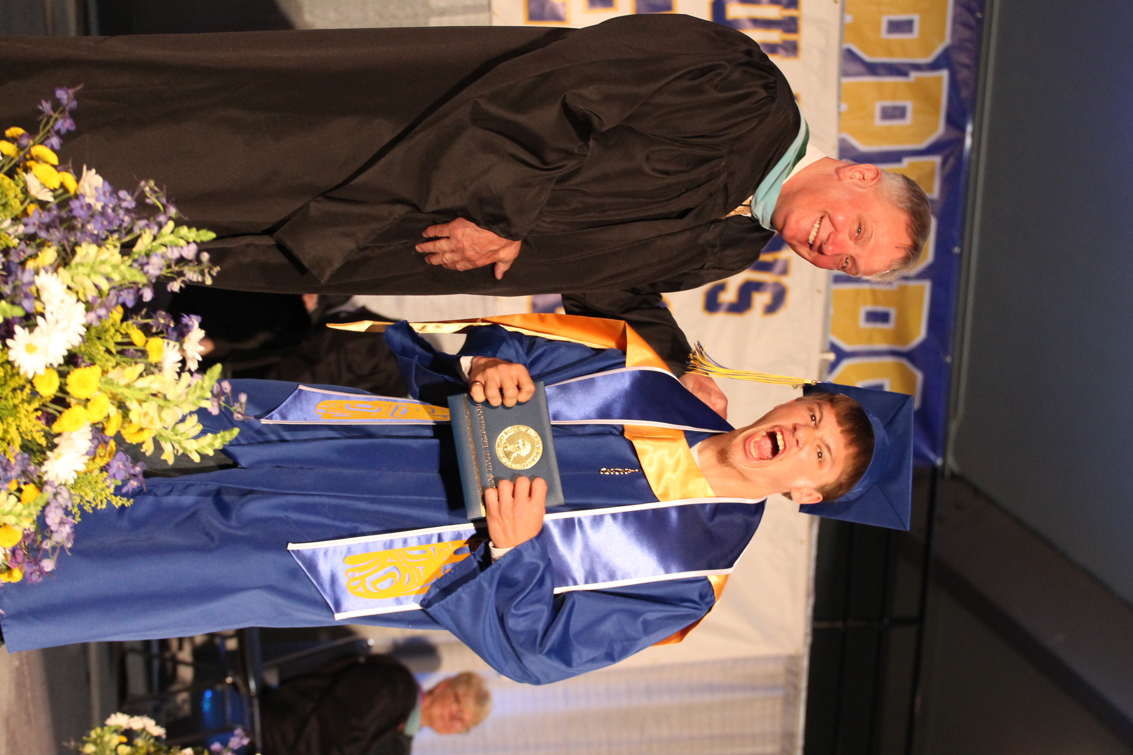 A male student in a blue graduation gown stands next to a faculty member, both smiling, on a stage with flowers in the foreground.