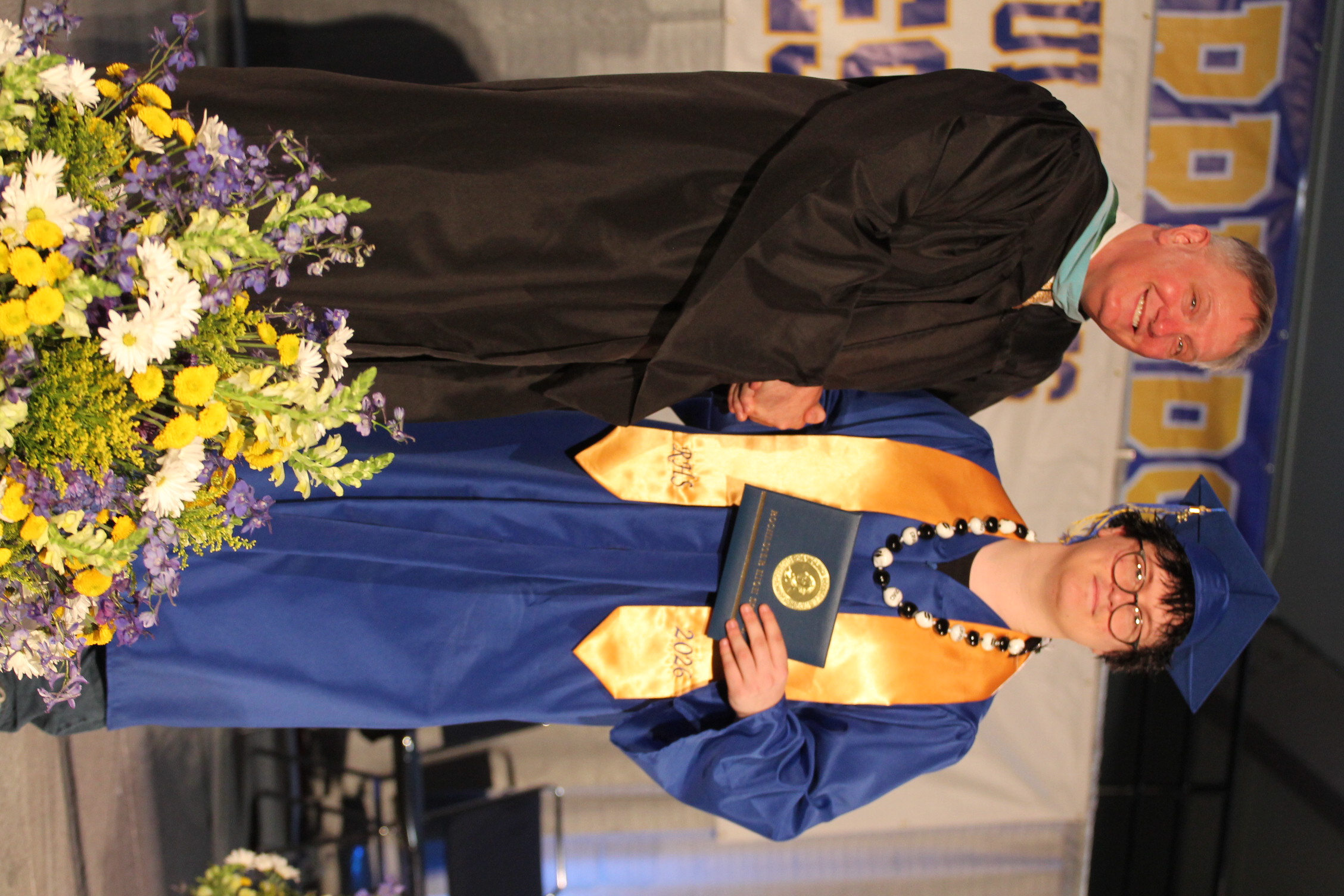 A person in a graduation gown and cap stands next to a man in a black gown.