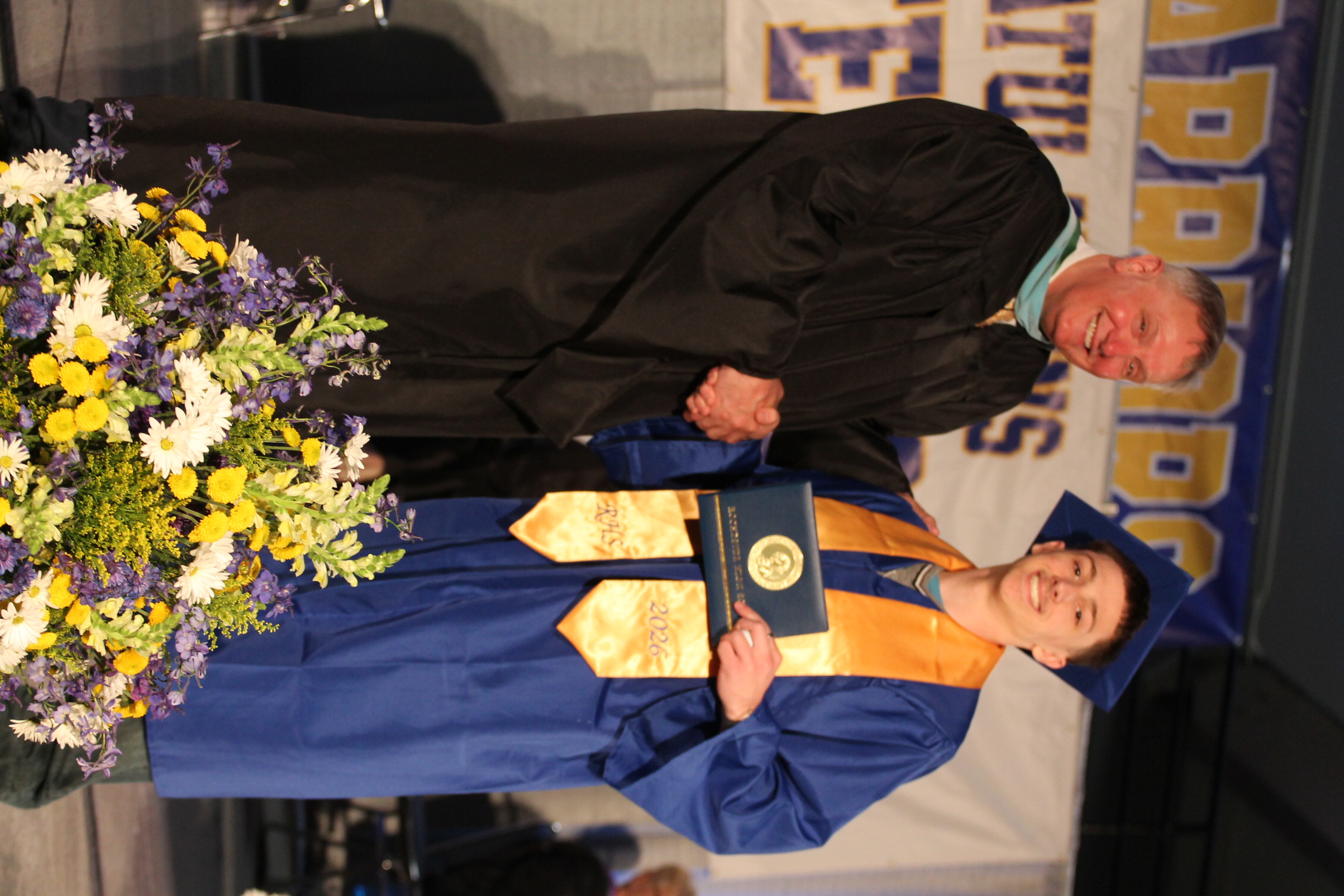A graduate in a blue gown and cap shakes hands with a faculty member in a black gown. They stand on a stage with flowers.