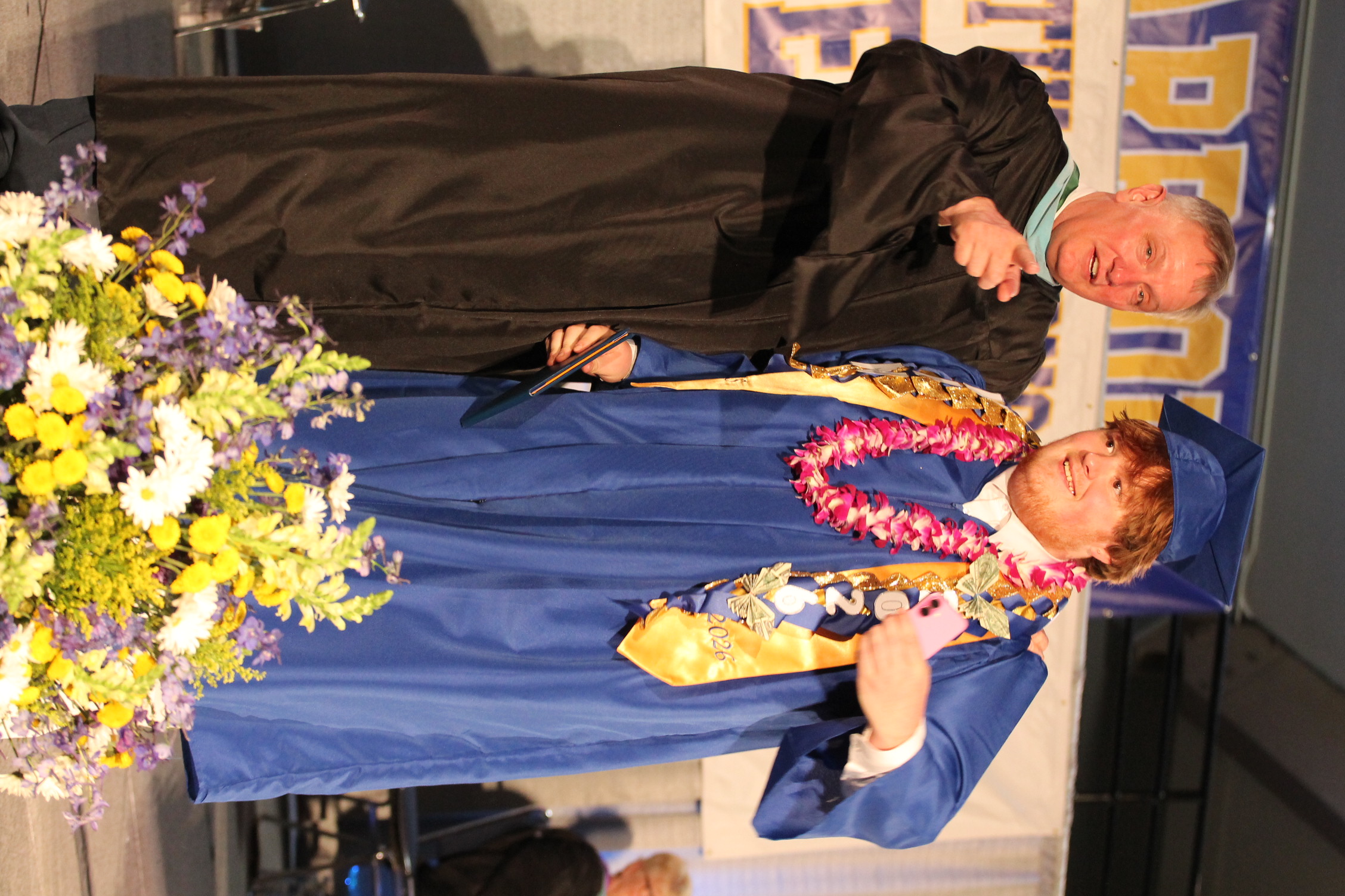 Two men stand on stage with a flower arrangement. One wears a black gown, the other a blue gown with a lei.