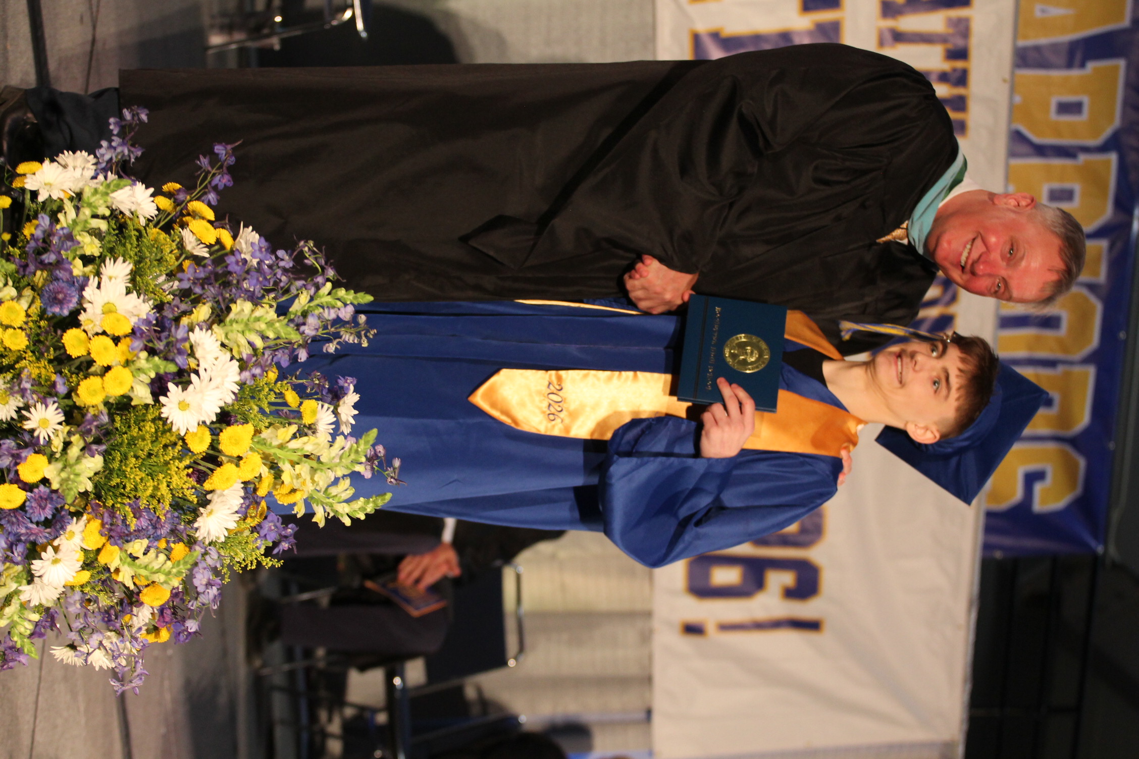 A male teacher and a male student in graduation gowns on a stage. The student holds a diploma.