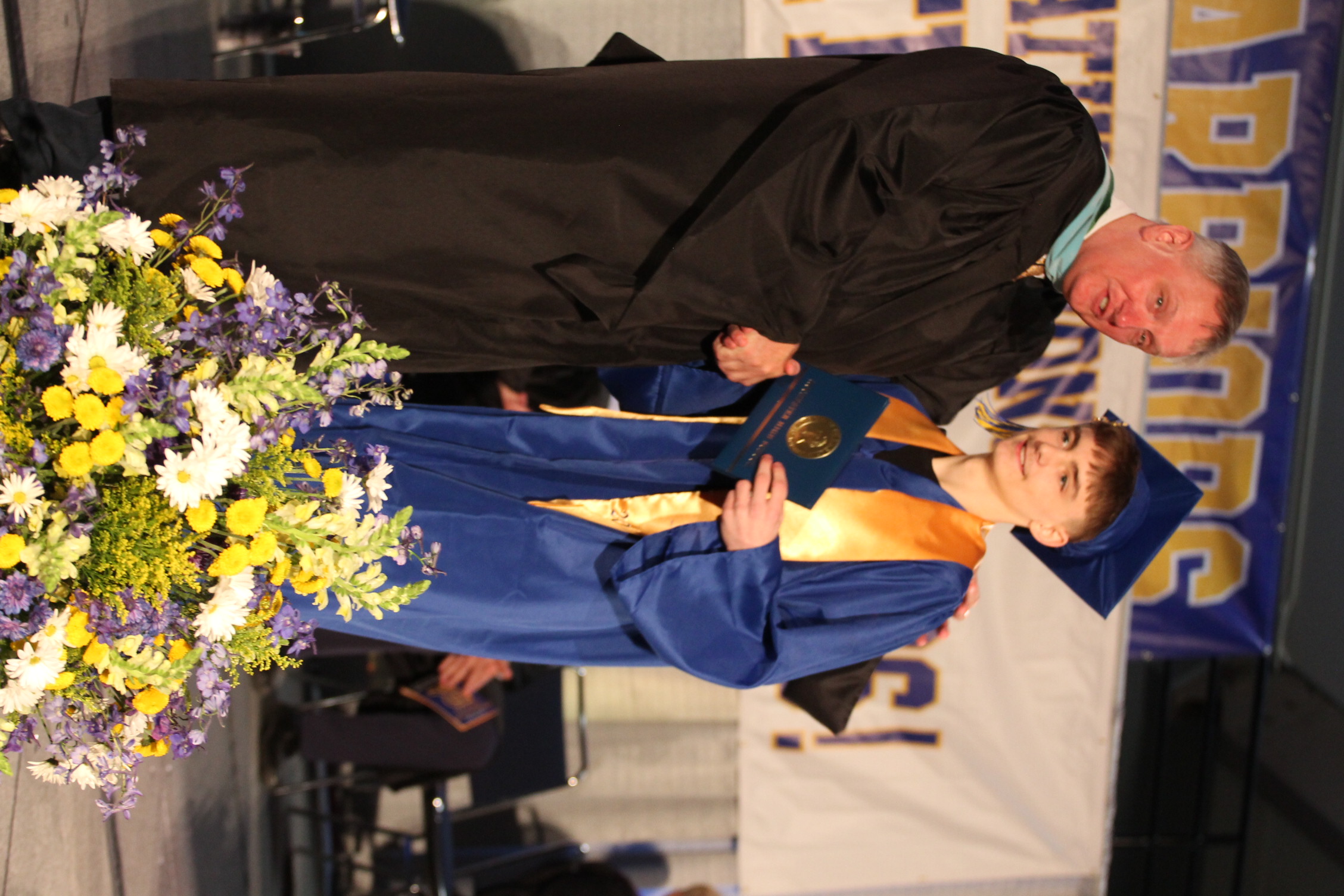 A man in black graduation robes gives a diploma to a young man in blue robes. Behind them is a large banner.