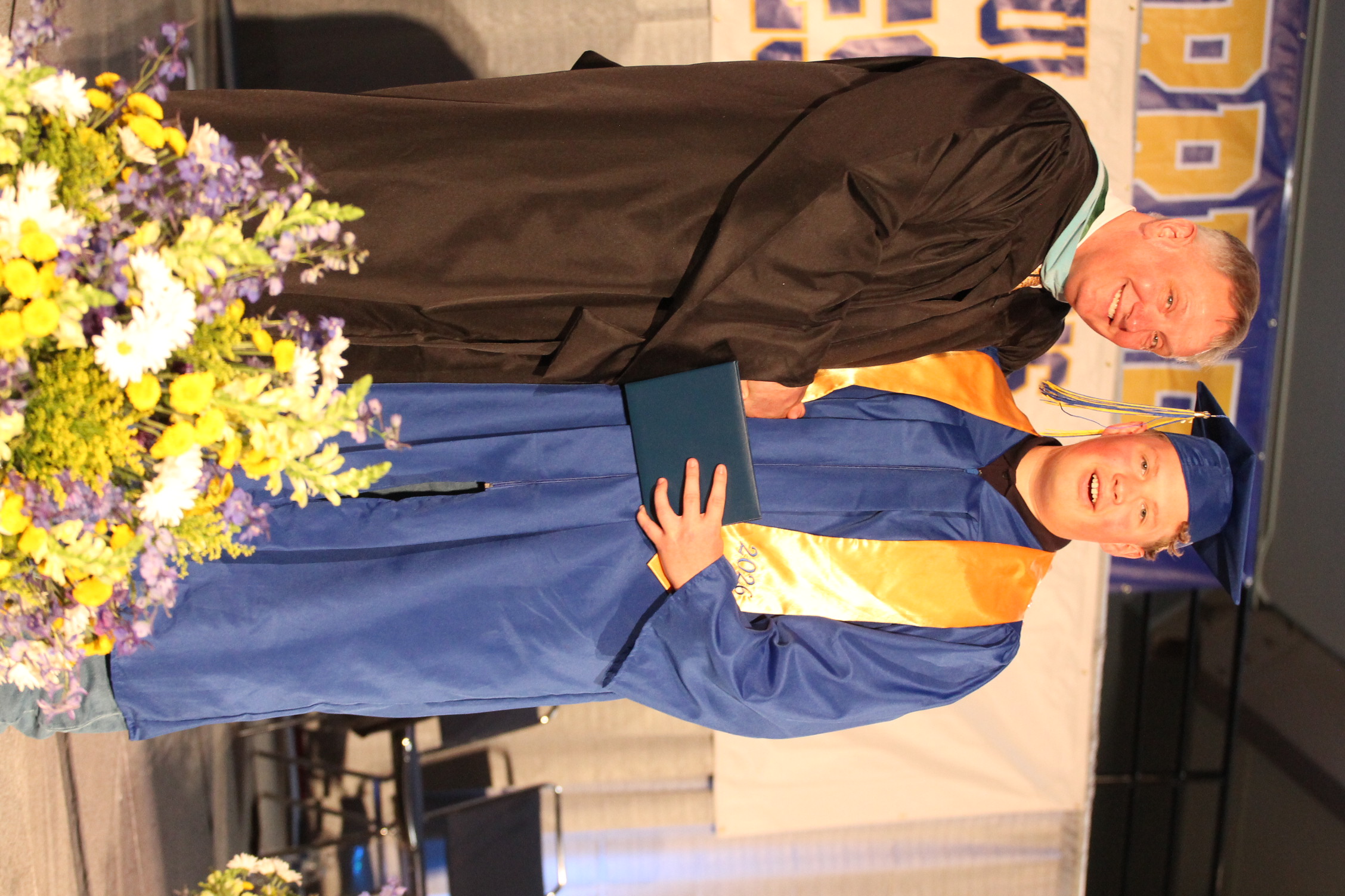 Man in black robe and cap shakes hands with graduate in blue gown and cap. Flower arrangement in foreground.