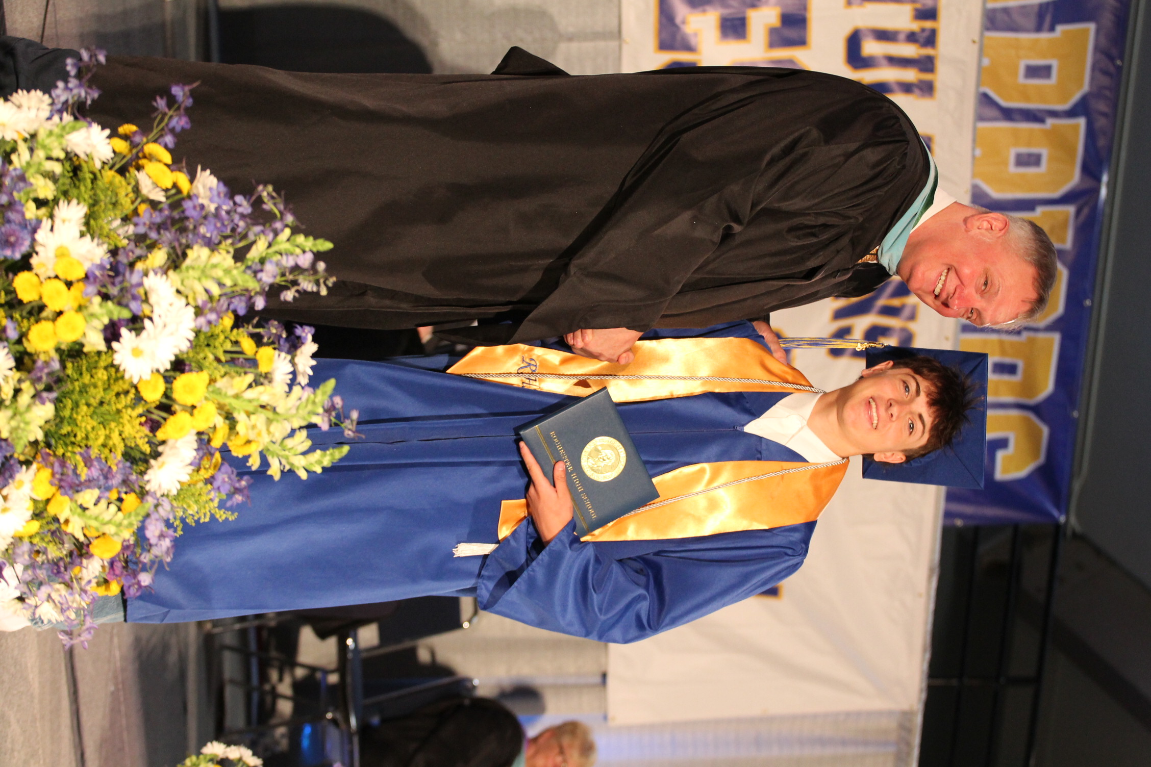 A man in a black robe shakes hands with a young graduate wearing a blue gown and a cap.