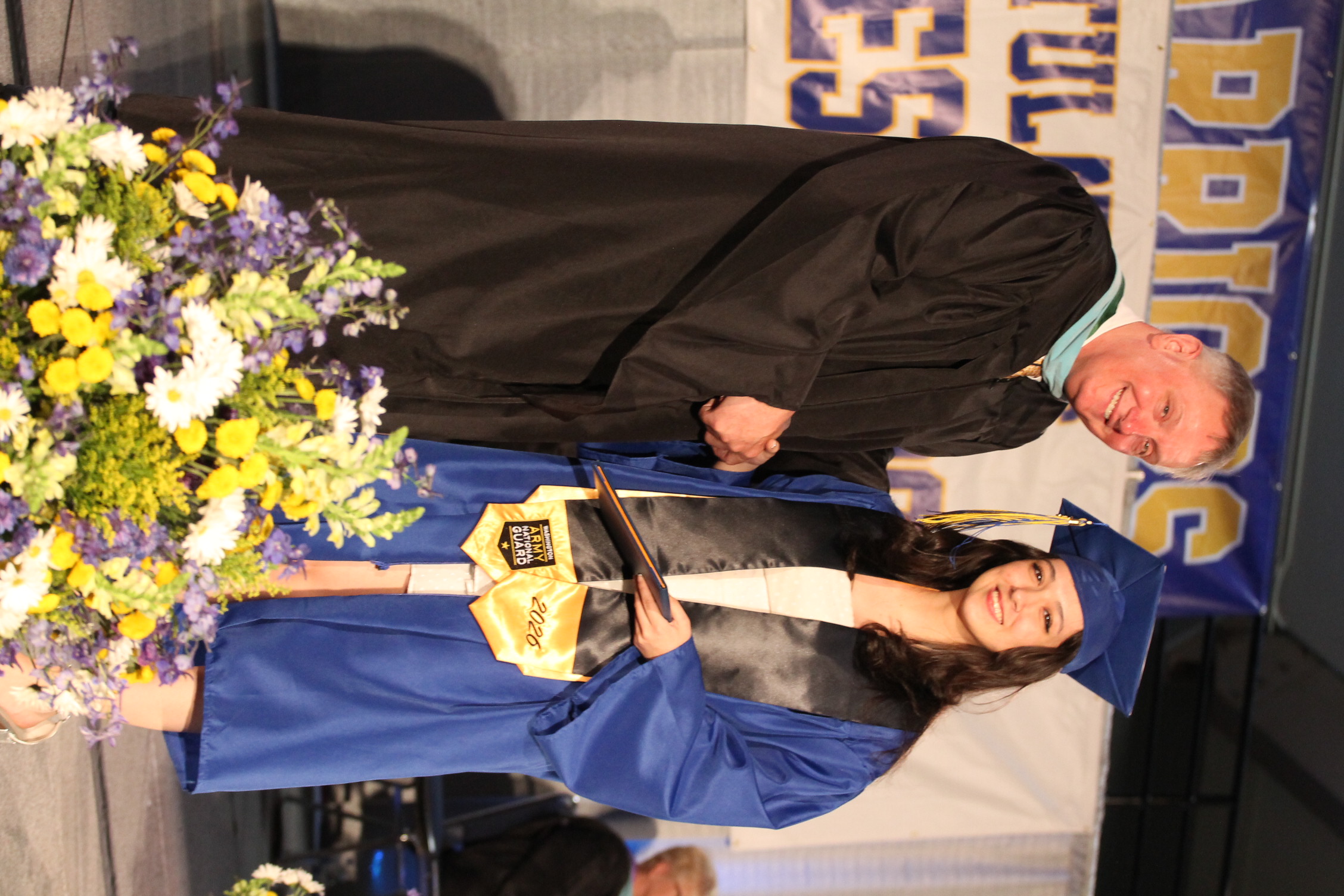 A man in a black graduation gown shakes hands with a woman in a blue gown. She holds a diploma. Flowers are in the foreground.
