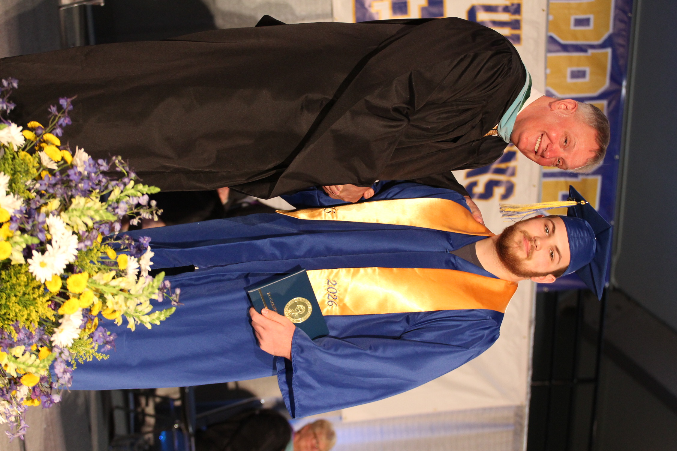 Two men in graduation robes stand on stage; one holds a diploma, and the other shakes hands. A flower arrangement is in the foreground.