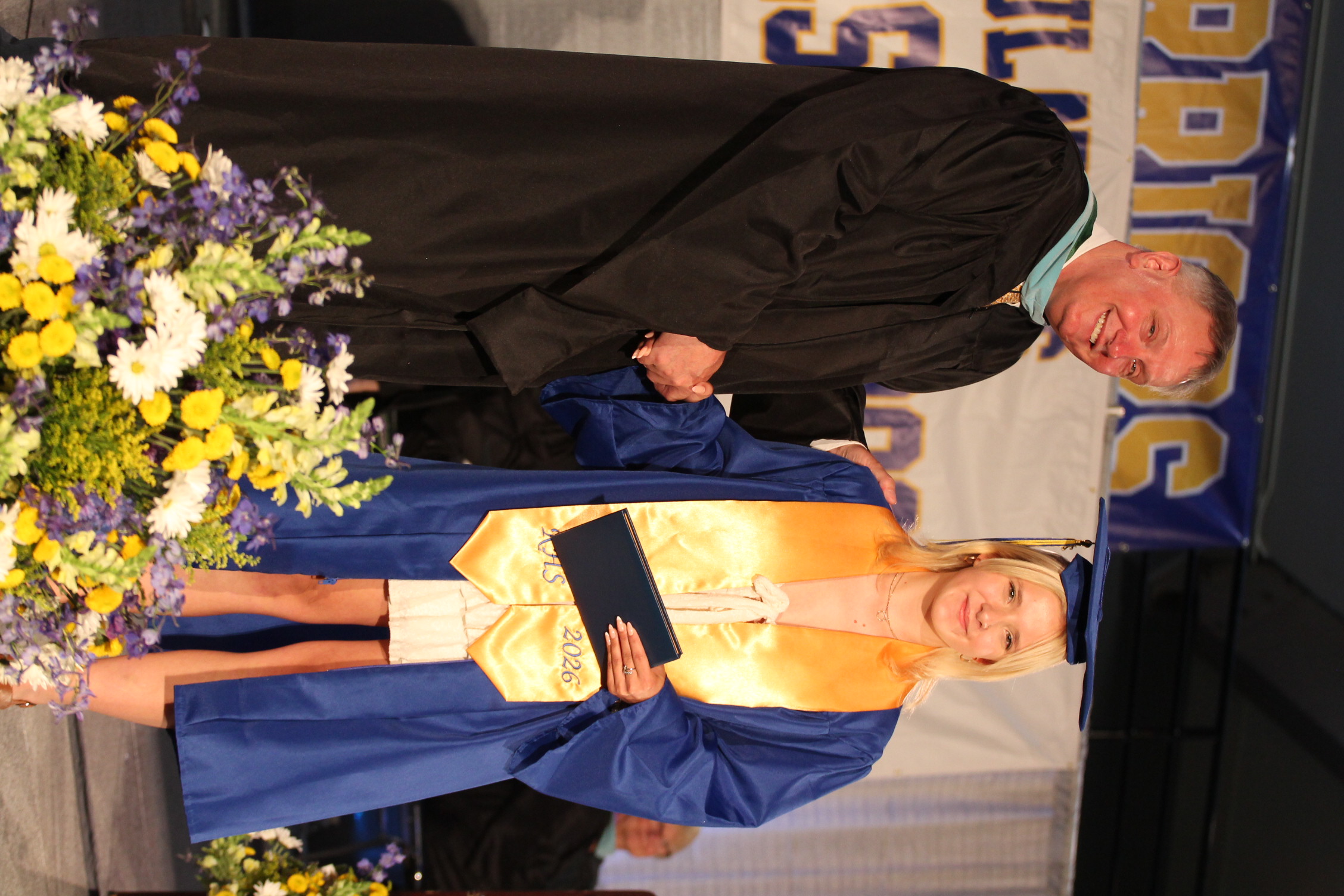 A man and a woman in graduation robes, the man is shaking the woman's hand. They are standing on stage.