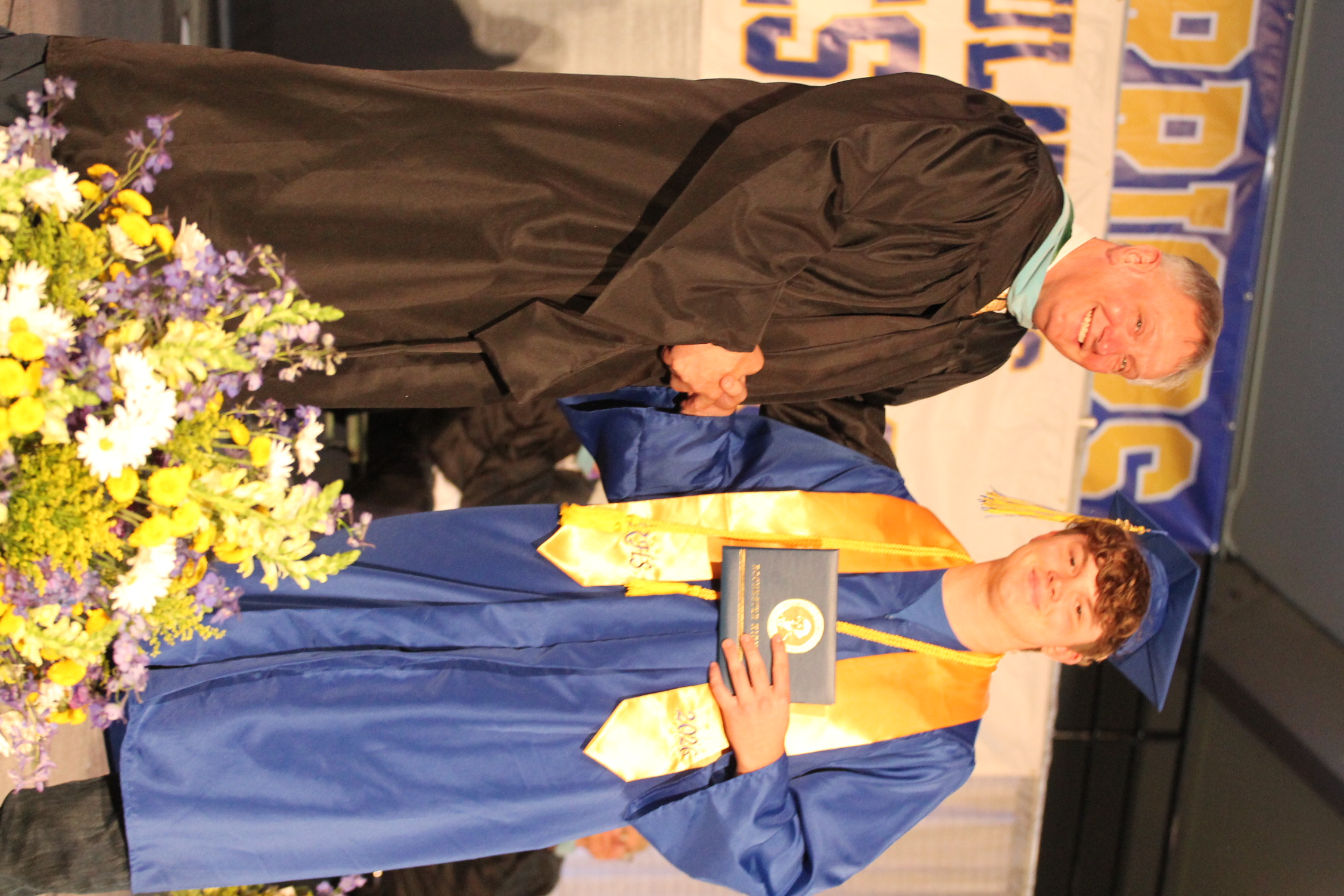 A man in a black gown shakes hands with a young man in a blue gown and cap. They stand near flowers on stage.