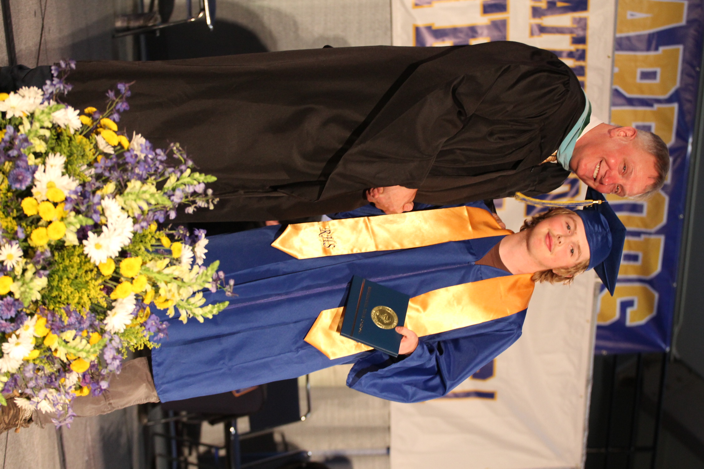 A man in a black gown and a woman in a blue gown and cap stand on stage. They hold a diploma.
