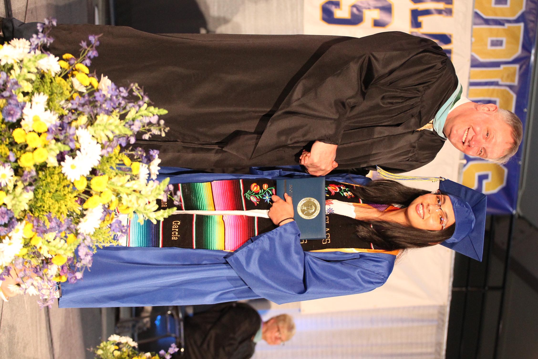 A man and woman in graduation robes exchange a handshake. The woman holds a diploma. Behind them, a banner with "DING" and a person sits on the stairs.