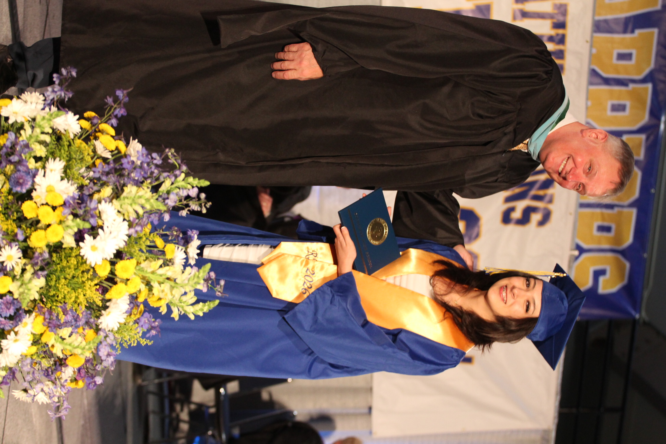 A man in a black gown hands a diploma to a woman in a blue gown at a graduation ceremony.